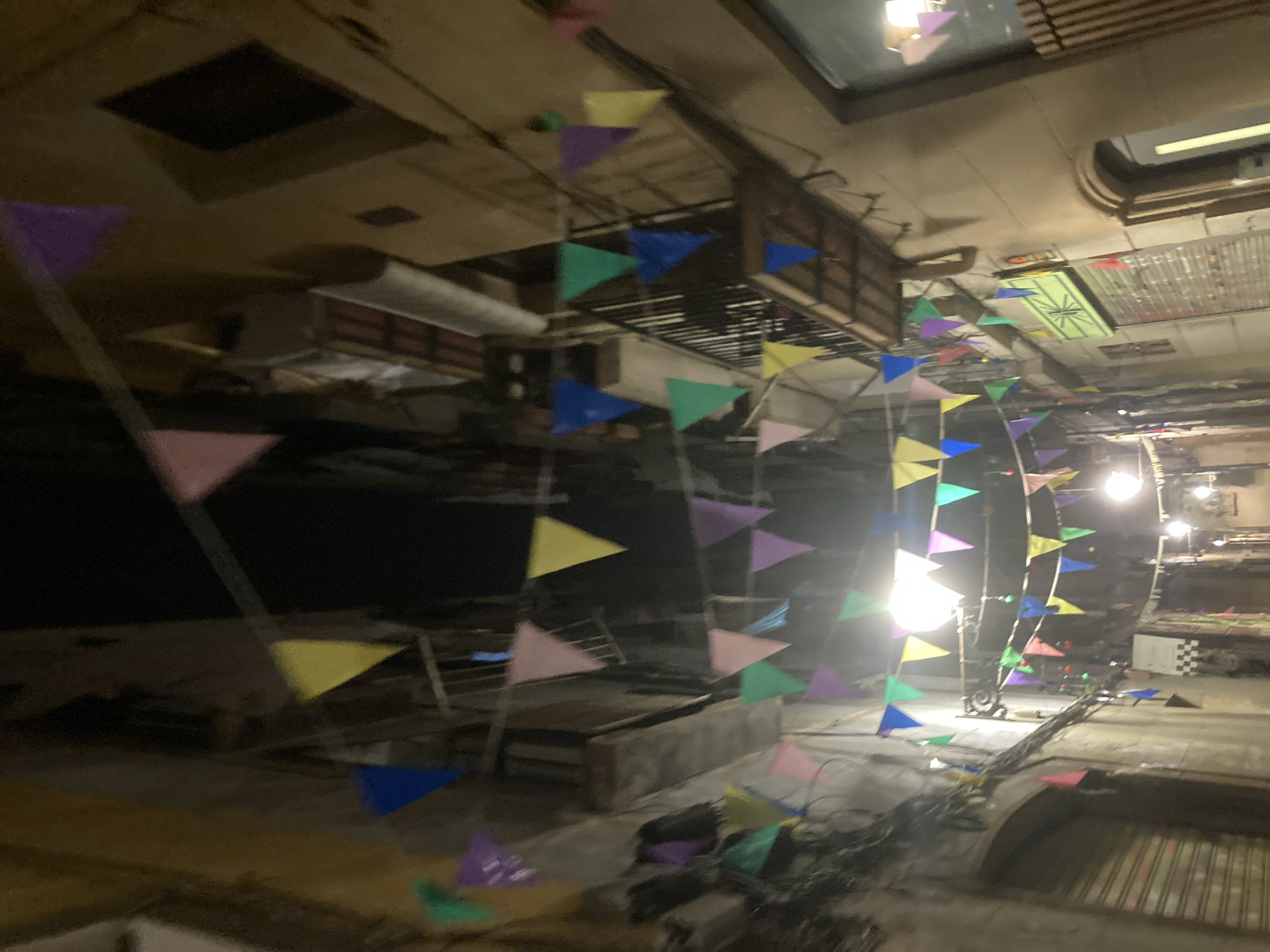 The image shows a narrow street in Barcelona, Spain, decorated with colorful triangular flags. The flags are strung across the street, creating a festive atmosphere. The buildings on either side of the street are tall and narrow, with balconies and shuttered windows. The street is dimly lit, suggesting that it is nighttime. There are no people visible in the image, but the flags suggest that an event or celebration is taking place.