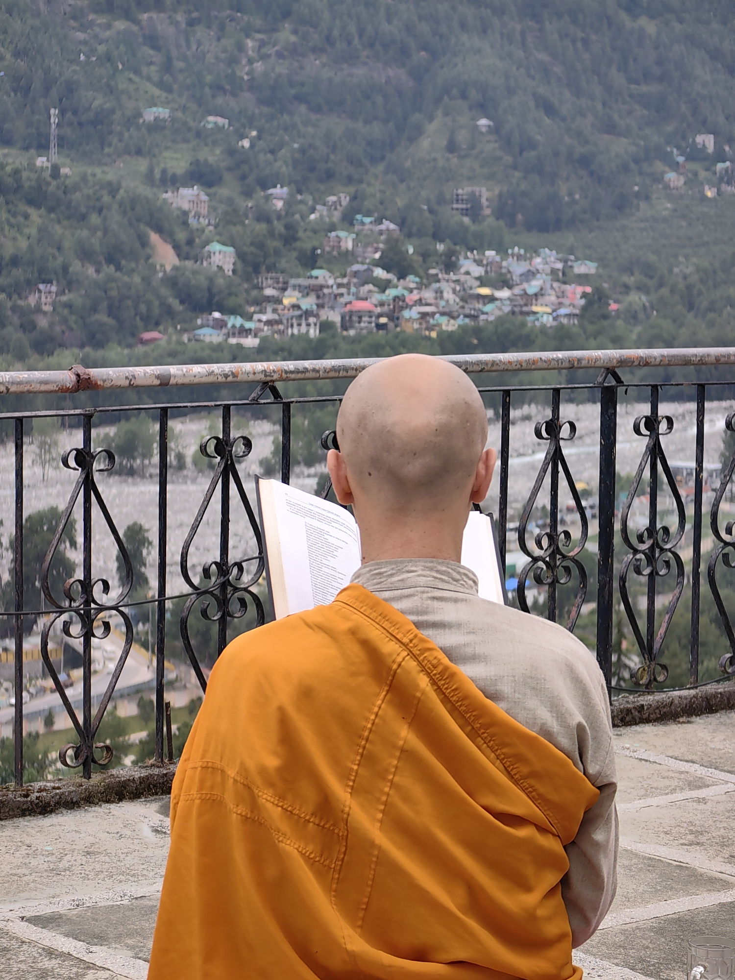 In Vashisht, India, an individual with a shaved head, wearing a beige robe and an orange saffron cloak, is seated on a stone patio, facing away from the camera. They are holding an open book and appear to be reading. The person is positioned in front of a black wrought-iron railing, behind which a blurred landscape of a hillside town dotted with buildings and trees unfolds. The railing has decorative scrollwork. The overall scene is captured from a medium distance, with the background slightly out of focus.
