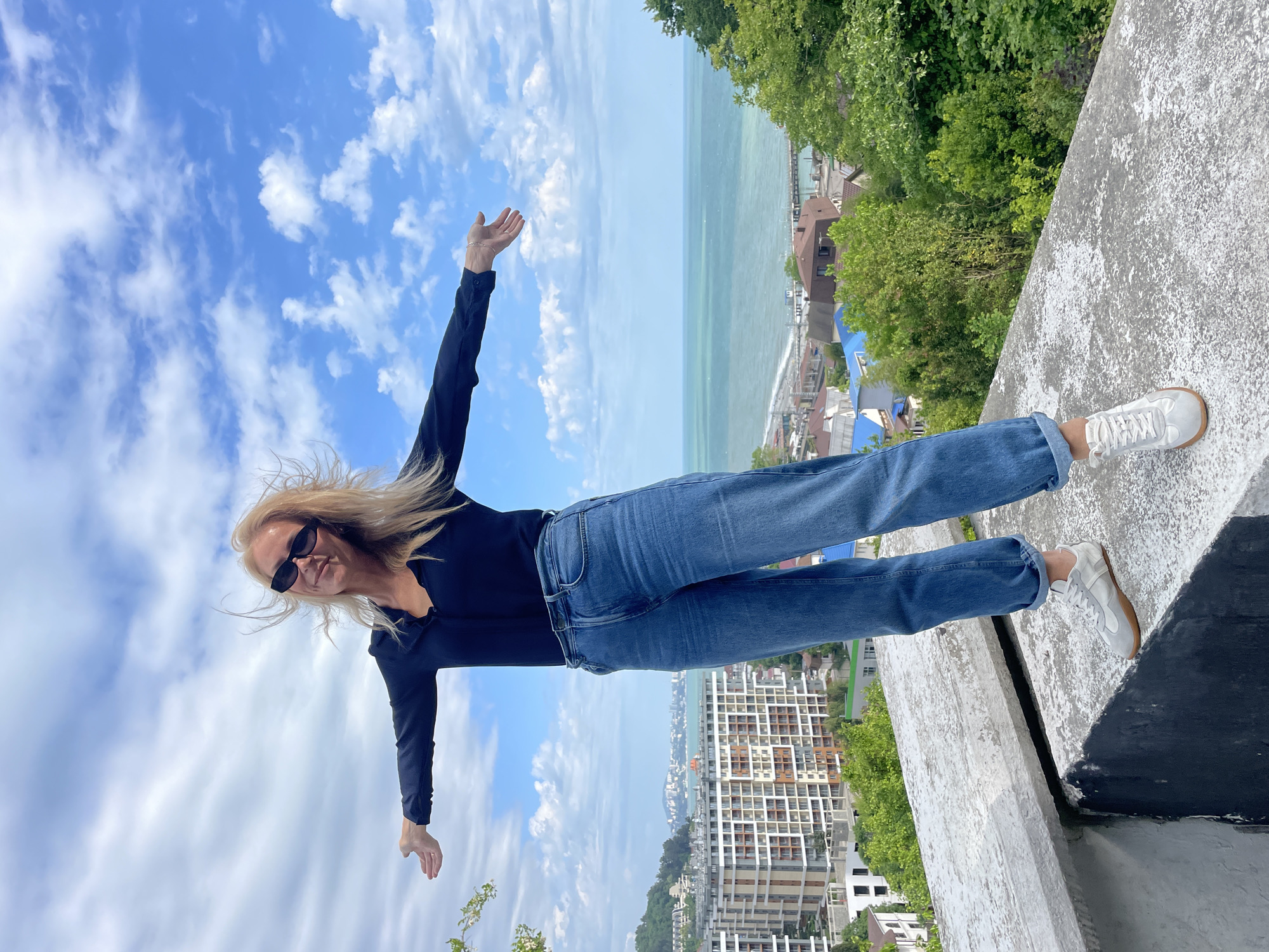 A woman with blonde hair and sunglasses stands on a ledge overlooking a cityscape, the Black Sea, and the sky in Dagomys, Russia. She wears a black top and blue jeans, and her arms are outstretched. The backdrop includes a blue sky with clouds, the sea, and urban buildings. A concrete ledge is under her feet.