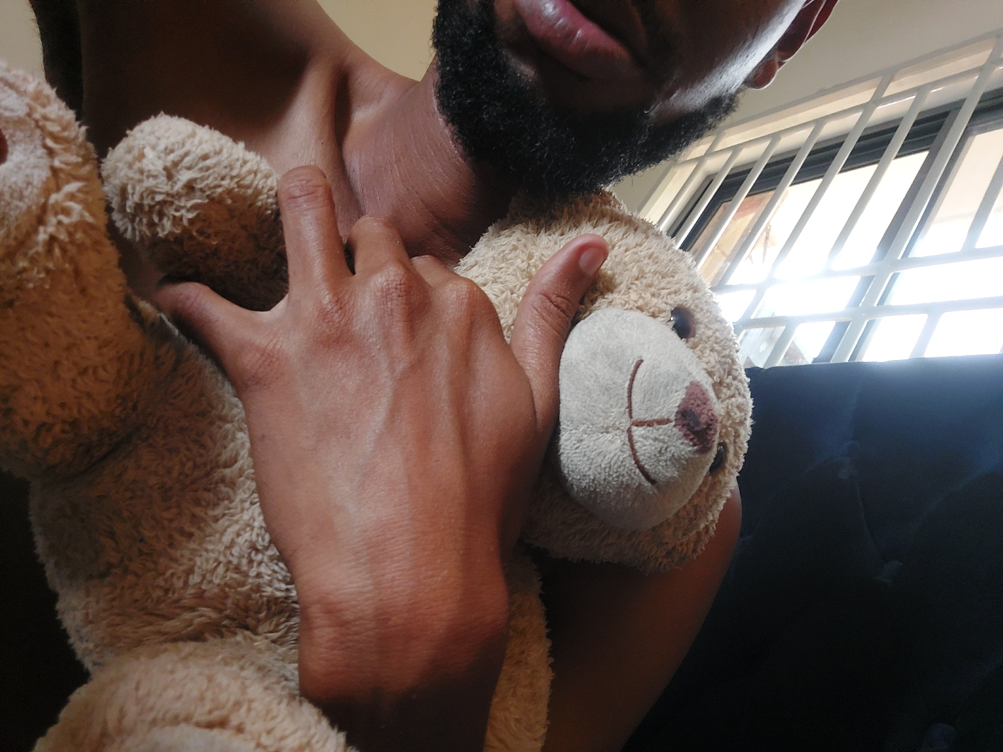 A shirtless man with a dark beard is seen holding a beige teddy bear. The man's bare chest and part of his face are visible. The teddy bear has a light-colored snout with stitched details representing the nose and mouth. Light is entering from a window with what appears to be metal bars. The presence of metal bars, the type of window, or other location context in the image DO NOT suggest this scene is located in the city of Dar es-Salaam, Tanzania.
