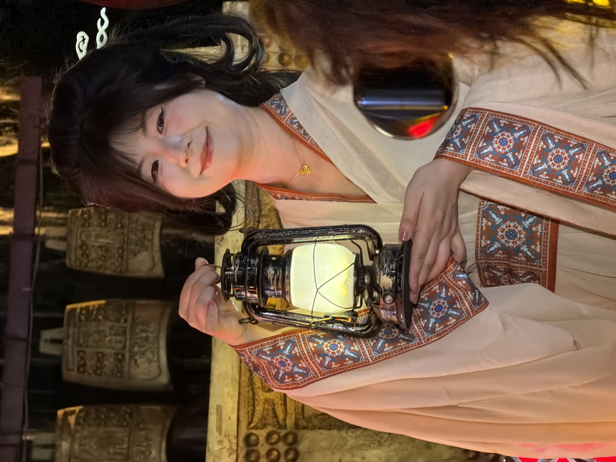 In the city of Yuzhong District, China, a woman in traditional attire holds a lantern. The woman has dark hair styled up and is smiling at the camera. She is wearing a light-colored robe with decorative borders on the sleeves and front. The lantern is old-fashioned, made of dark metal with a glass enclosure that emits a warm light. Behind her, there are several large, ornate bells hanging, suggesting a cultural or historical setting. The lighting in the scene is dim, with the lantern providing a significant source of illumination.
