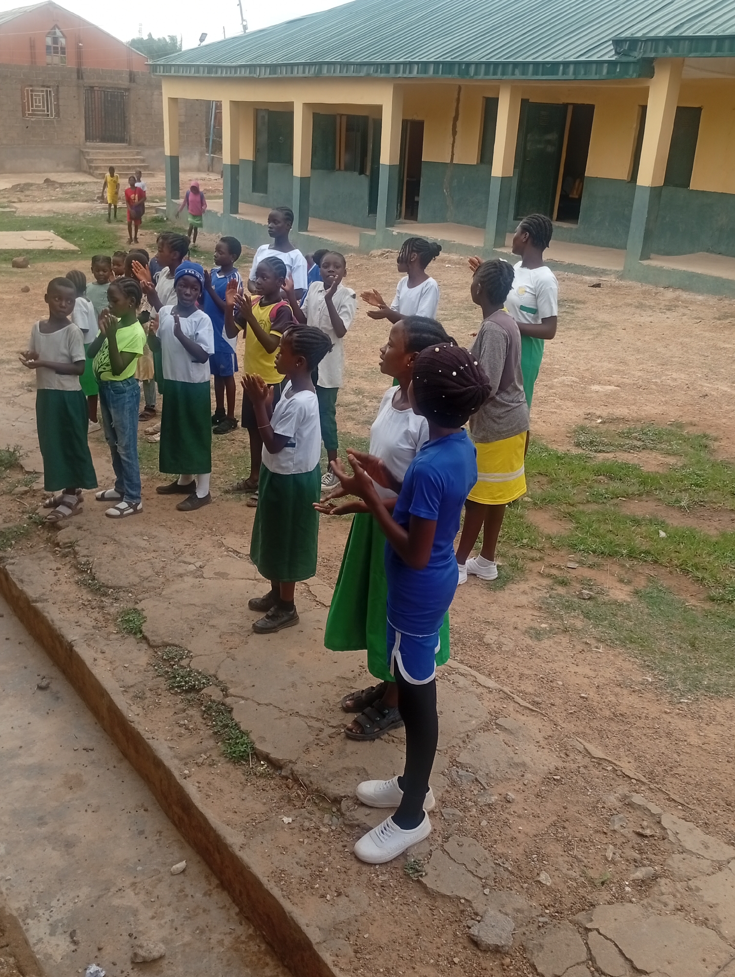 A group of children, primarily girls, are standing outdoors, most with their hands clasped together. They appear to be in a schoolyard in Narayi, Nigeria, evidenced by the school building in the background. The children are wearing various clothes, some with green skirts and white shirts, others in more casual attire like jeans and t-shirts, or athletic wear. Some children in the background are walking or running. The ground is a mix of concrete and dirt, with some patches of grass. A building with a green roof is visible in the background.