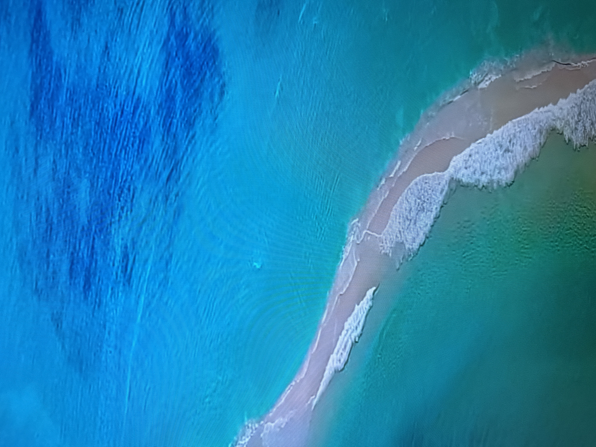 This aerial view depicts a narrow strip of sandy beach with turquoise and deep blue water on either side. Gentle waves are breaking onto the shore, creating a foamy white edge. The water exhibits variations in color, transitioning from a vibrant turquoise near the beach to a darker, richer blue in the distance. The texture of the water suggests some currents or underwater topography, particularly in the deeper blue area. There are no visible people or man-made objects. The scene is natural, likely a coastal environment. The time of day appears to be daytime, with good visibility. No text is visible in the image. The location context of Almaty, Kazakhstan, is not directly supported by visual cues in this image, as it depicts a coastal scene, and Almaty is a landlocked city.