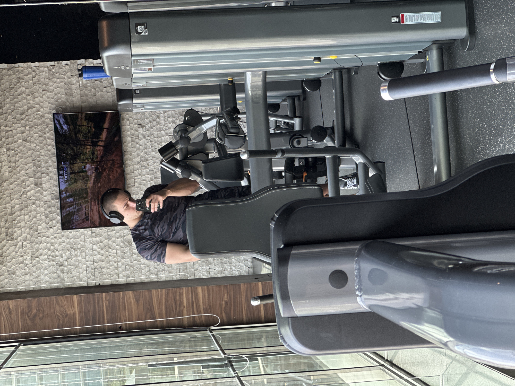 The image depicts a modern gym or fitness facility, likely located in Sofia, Bulgaria, during the daytime, suggested by the bright natural light.
The primary subject is a man seated on a grey and black multi-station gym machine. He has short dark hair, is wearing black over-ear headphones, a dark camouflage-patterned t-shirt, and dark shorts. He is actively drinking from a dark-colored water bottle or cup. Part of another gym machine, possibly a treadmill, is visible in the foreground.
The setting features a wall covered in light-colored, textured stone or tile behind the man. A large flat-screen television is mounted on this wall, displaying a serene forest scene with the text "THEME ADVENTURE TO YOUR RHYTHM" partially visible. The floor is a dark grey, textured surface, indicative of gym flooring. A wide window or glass partition extends across the lower portion of the image, offering a view of the outside or an adjacent indoor area, with wooden paneling underneath. White beaded chains for window treatments are also visible.
A blue cylindrical object rests on a ledge behind the gym equipment. The gym machine itself has visible branding, "PRECOR," and a warning label with unreadable text.
The man appears to be taking a break from his workout, hydrating and listening to something via his headphones, creating a relaxed yet engaged atmosphere within the well-equipped and brightly lit fitness space.