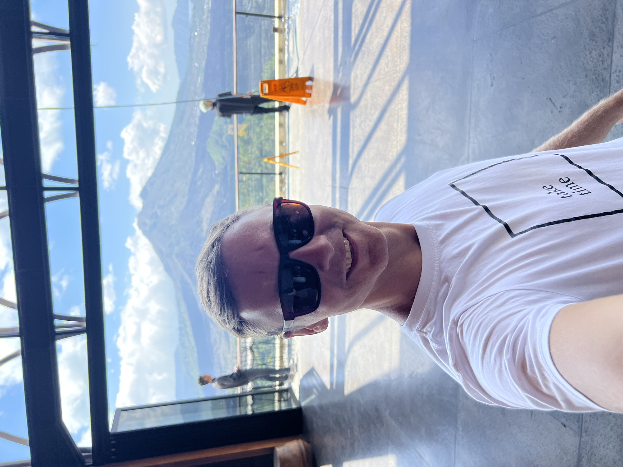 In Kintamani, Indonesia, a man wearing a white t-shirt and sunglasses takes a selfie in front of a scenic view of a mountain and lake. The man is positioned in the foreground, smiling at the camera. In the background, two figures stand near a railing, looking out at the landscape. One figure is on the left, dressed in a gray shirt and dark pants, while the other is on the right, wearing a black shirt and pants. An orange caution sign is visible on the right side of the frame. The environment appears to be an elevated viewing area with large windows and a balcony, offering a panoramic vista of the natural scenery, including a large mountain peak in the distance under a partly cloudy sky. The floor is tiled, and sunlight casts shadows.