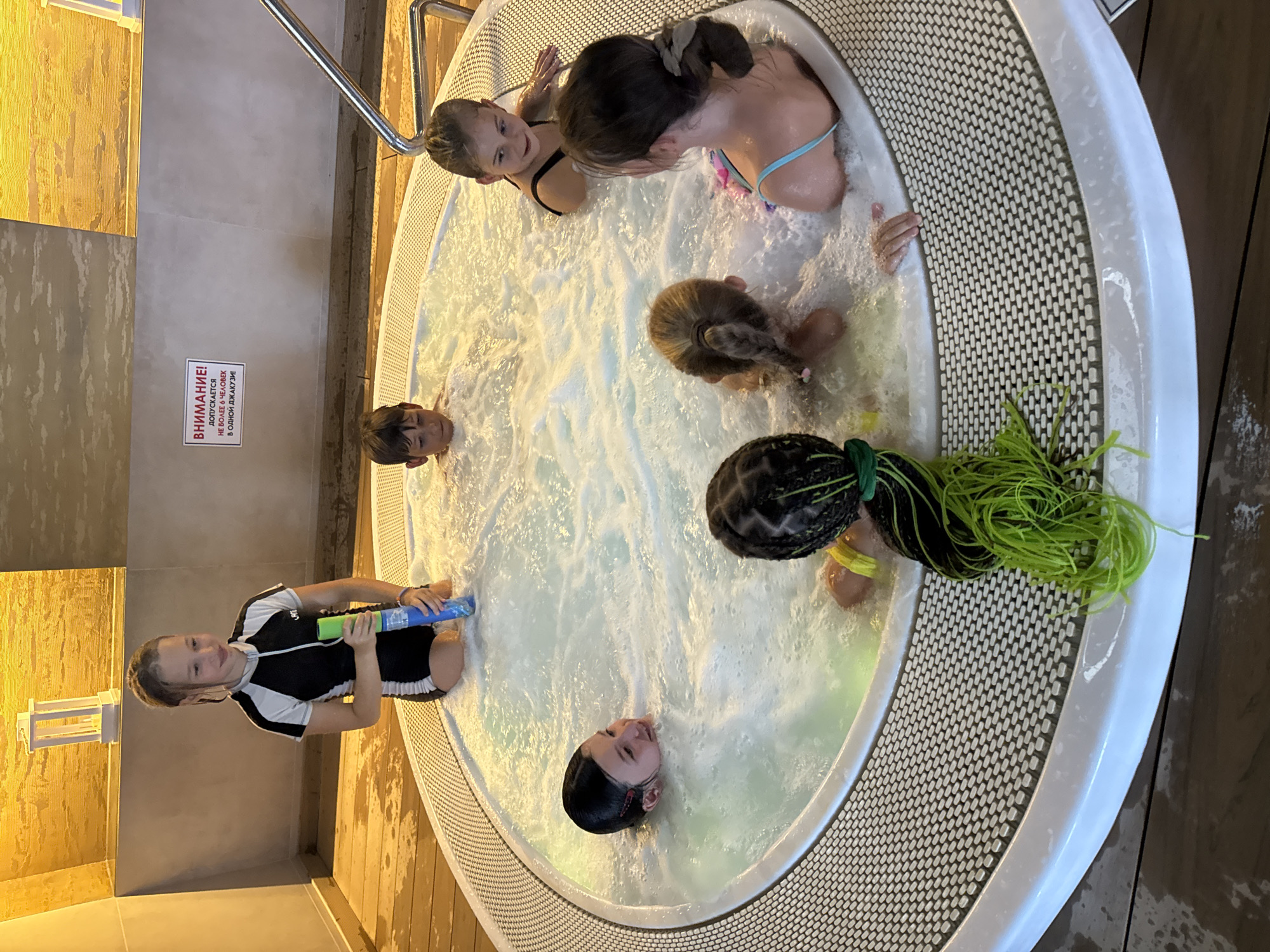 In Yevpatoriya, Ukraine, a group of seven children are in a hot tub. The children, ranging in age from approximately 5 to 12 years old, are wearing swimsuits and are immersed in bubbly, heated water. One girl in the foreground, wearing a black and white swimsuit, kneels at the edge of the tub, holding a green and blue water toy. Another girl with long, bright green braided hair is visible in the lower part of the frame, facing away from the camera. Several other children are visible in the water, some smiling and looking at the camera. The hot tub is circular with a textured white rim and is situated on a wooden floor. In the background, a sign with Russian text is mounted on a wall, and a white decorative lantern is visible on a shelf. The lighting in the room appears warm and artificial.