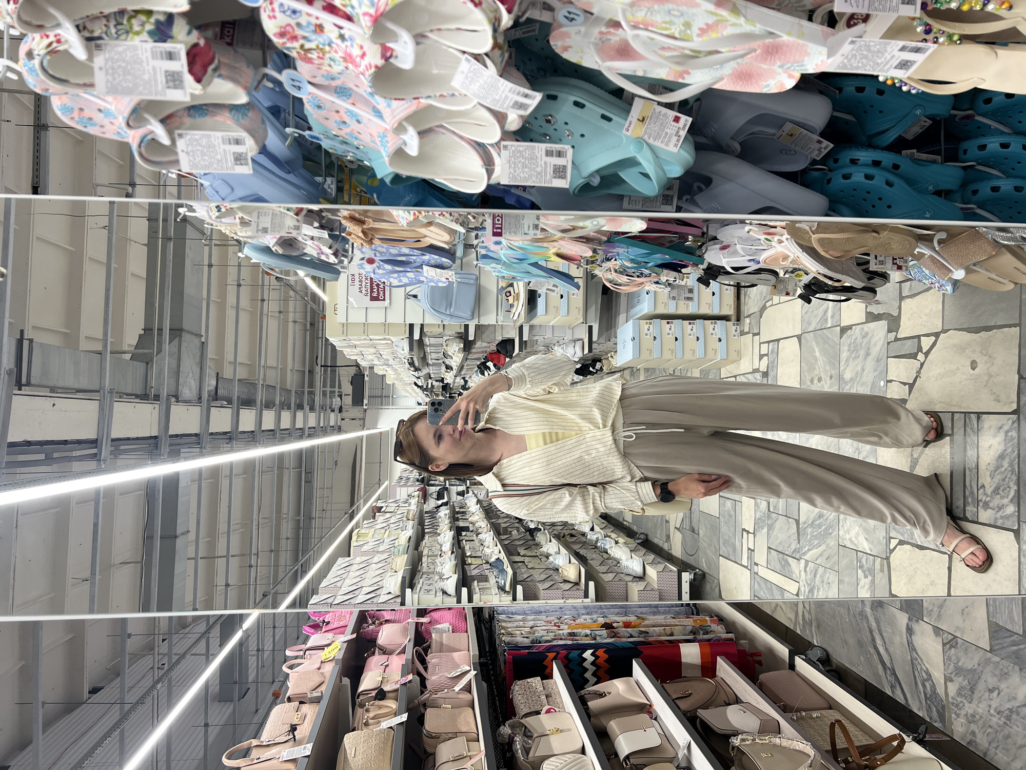 In the city of Chaikovsky, Russia, a young woman stands in a brightly lit retail store, reflected in a large mirror. She is taking a selfie with her mobile phone, wearing a light-colored, pinstriped open-neck shirt over a yellow top, and loose-fitting beige pants. Her outfit is casual, and she is wearing simple sandals on her feet. The store appears to be a department store or a large supermarket, with aisles dedicated to various merchandise. On her left, shelves are filled with an assortment of handbags in various shades of pink and beige. To her right, an aisle displays a wide selection of colorful flip-flops and clogs, hanging from racks. The flooring is a grey and white patterned tile. The background of the reflection shows more shelves stocked with products and the store's ceiling infrastructure, including fluorescent lighting and exposed conduits.