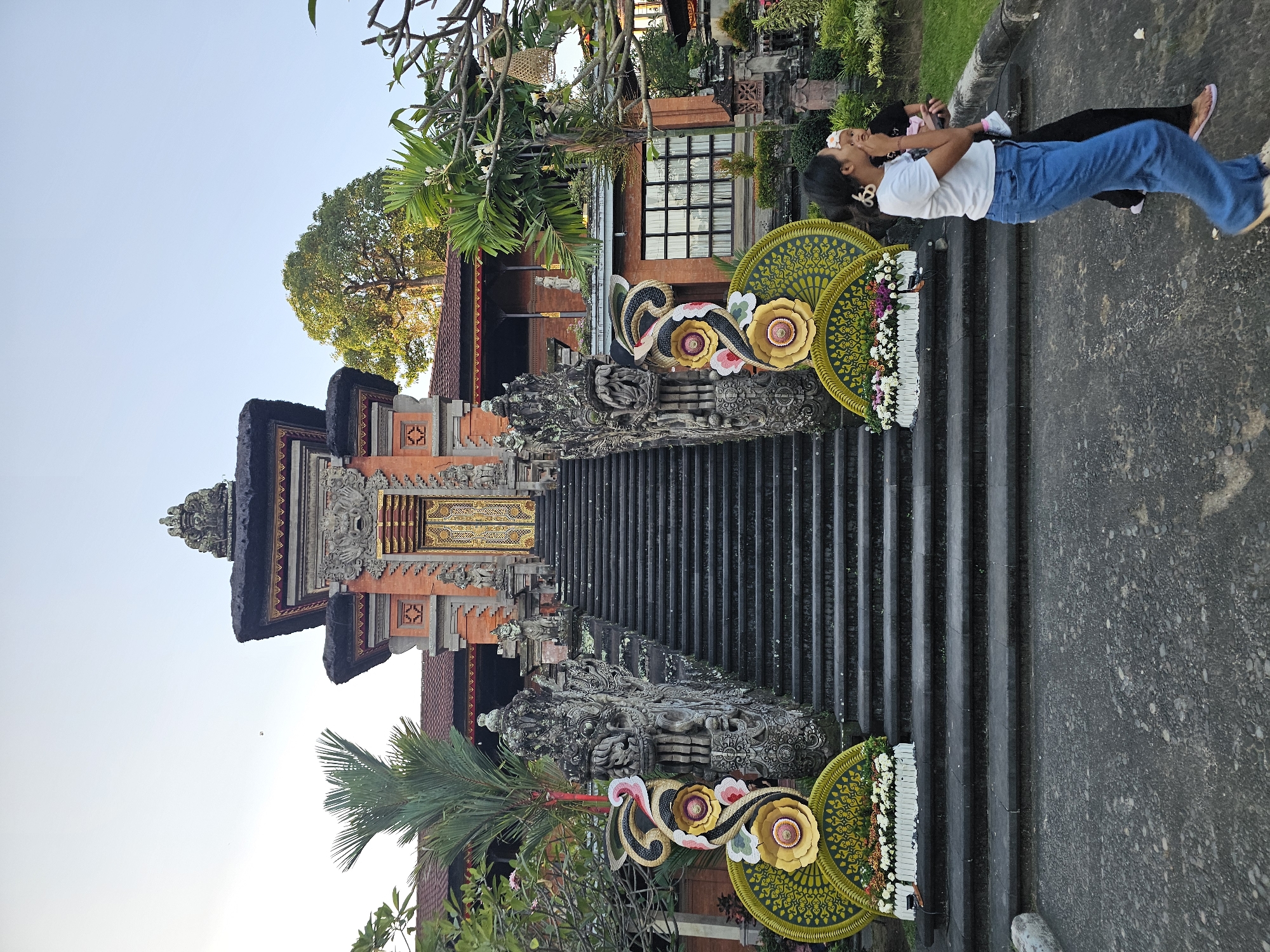 This is a medium shot, eye-level, outdoor photograph of a Balinese temple entrance. The temple features a grand, ornate staircase leading up to a large golden door. The staircase is flanked by detailed stone carvings of mythical creatures and decorative elements. Large, colorful floral arrangements in a fan shape are placed at the base of the stairs. Two women are walking in the foreground, with one carrying a baby. The temple is made of red brick and traditional thatched roofing. Lush green trees and vegetation surround the temple, with a clear blue sky overhead. The scene appears to be in Denpasar, Indonesia, given the architectural style.