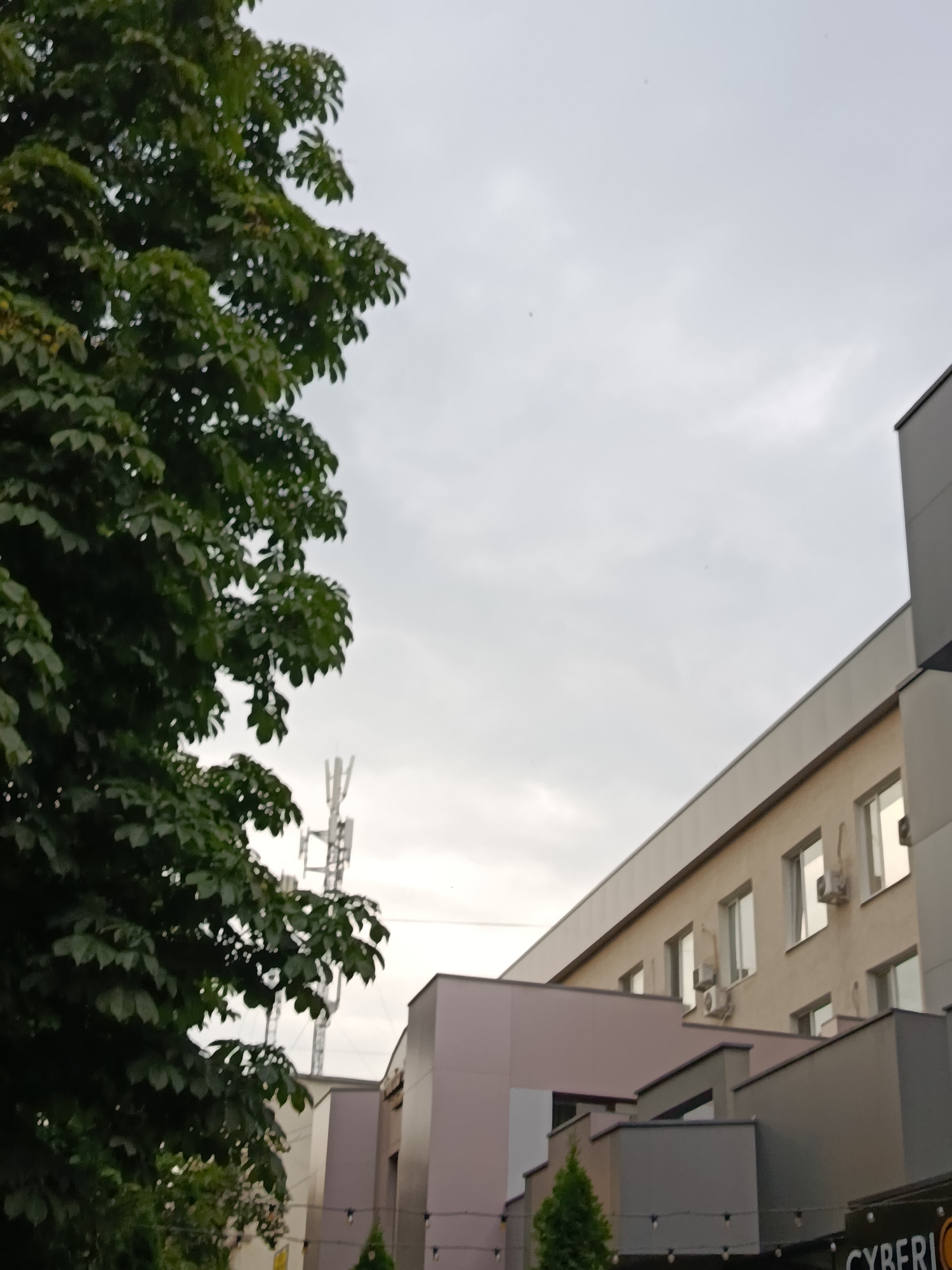 The image shows a modern building facade with multiple levels and windows, alongside a large, leafy tree. The architecture suggests an urban environment. The sky is overcast and light. A cell tower is visible in the background, to the left of the building. The building has several windows, some with air conditioning units attached. There are balconies or protruding sections on different levels of the building. The tree occupies the left side of the frame and its foliage is dense and green. The overall impression is a contemporary urban scene. The presence of a cell tower is a common feature in cities. The building's design, with its geometric shapes and multiple levels, is characteristic of modern urban architecture. The lighting suggests it is daytime, possibly late afternoon or morning due to the diffused light from the clouds.