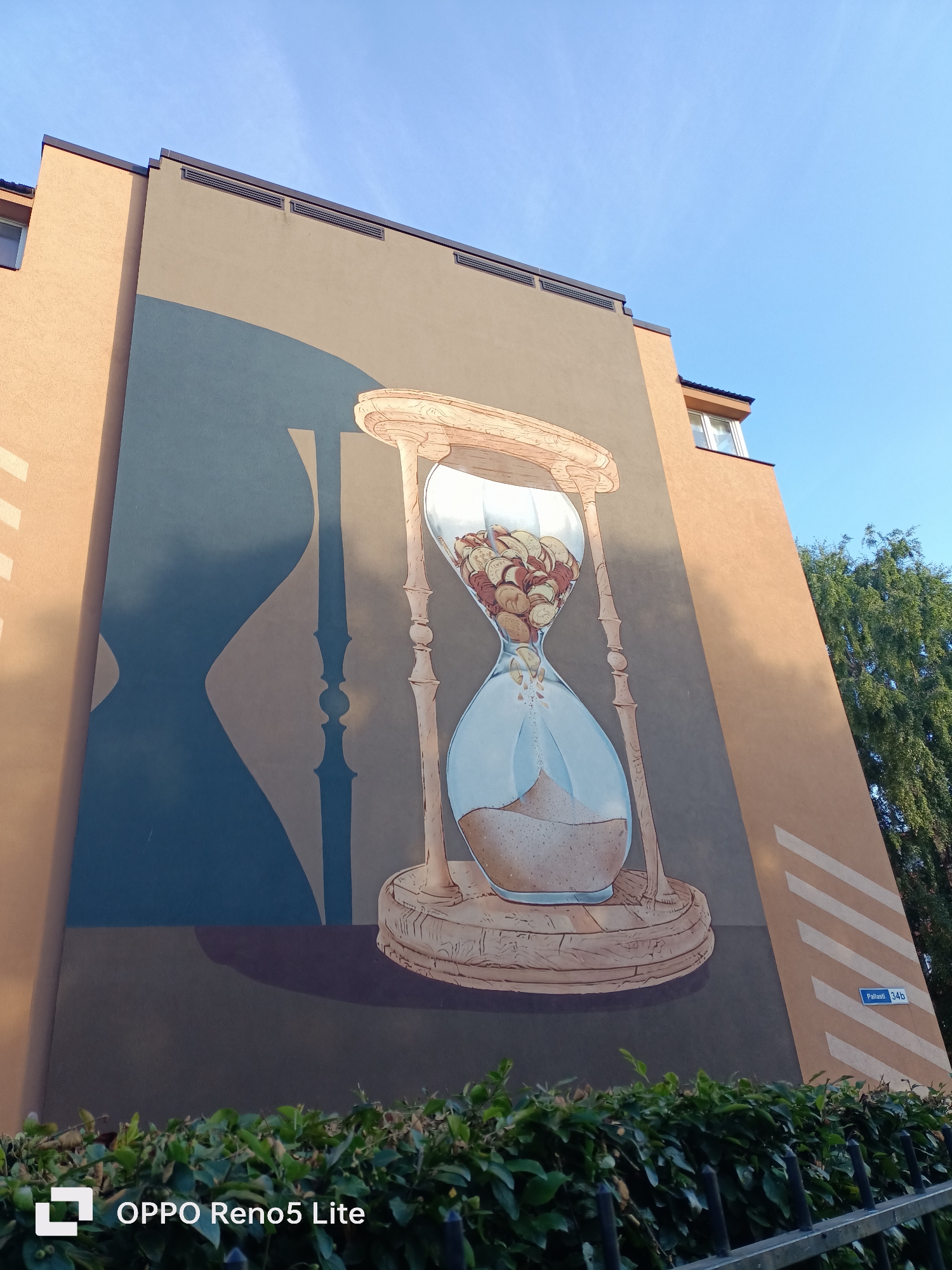 The image shows a mural on the side of a building in Tallinn, Estonia, depicting a large hourglass filled with coins. The mural covers a significant portion of the building's facade, which is painted in shades of brown and grey. The hourglass is rendered in a realistic style, with a wooden frame and a glass bulb containing numerous coins. A shadow of an hourglass is also visible on the left side of the mural. The building has windows and ventilation grilles, and a tree with green foliage is visible on the right side, along with a blue street sign that reads "Pallasti 34b". A hedge and a metal fence are in the foreground, partially obscuring the base of the building. The sky above is clear and blue. The bottom left corner of the image displays a watermark that reads "OPPO Reno5 Lite".