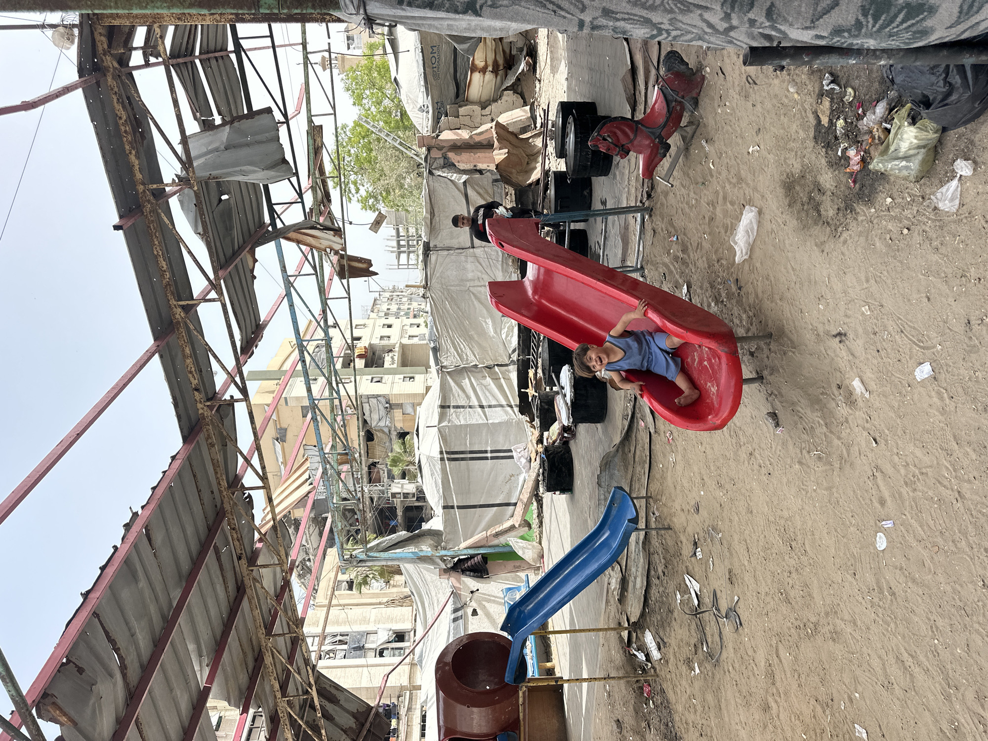 The photograph depicts a child playing on a red slide in a derelict area in Gaza City, Palestinian Territories. Overhead, a damaged metal roof structure suggests prior destruction. Background tents provide makeshift shelters. Another child is seen standing near the playground equipment. Rubbish is scattered on the sandy ground. Buildings in the backdrop show signs of damage.