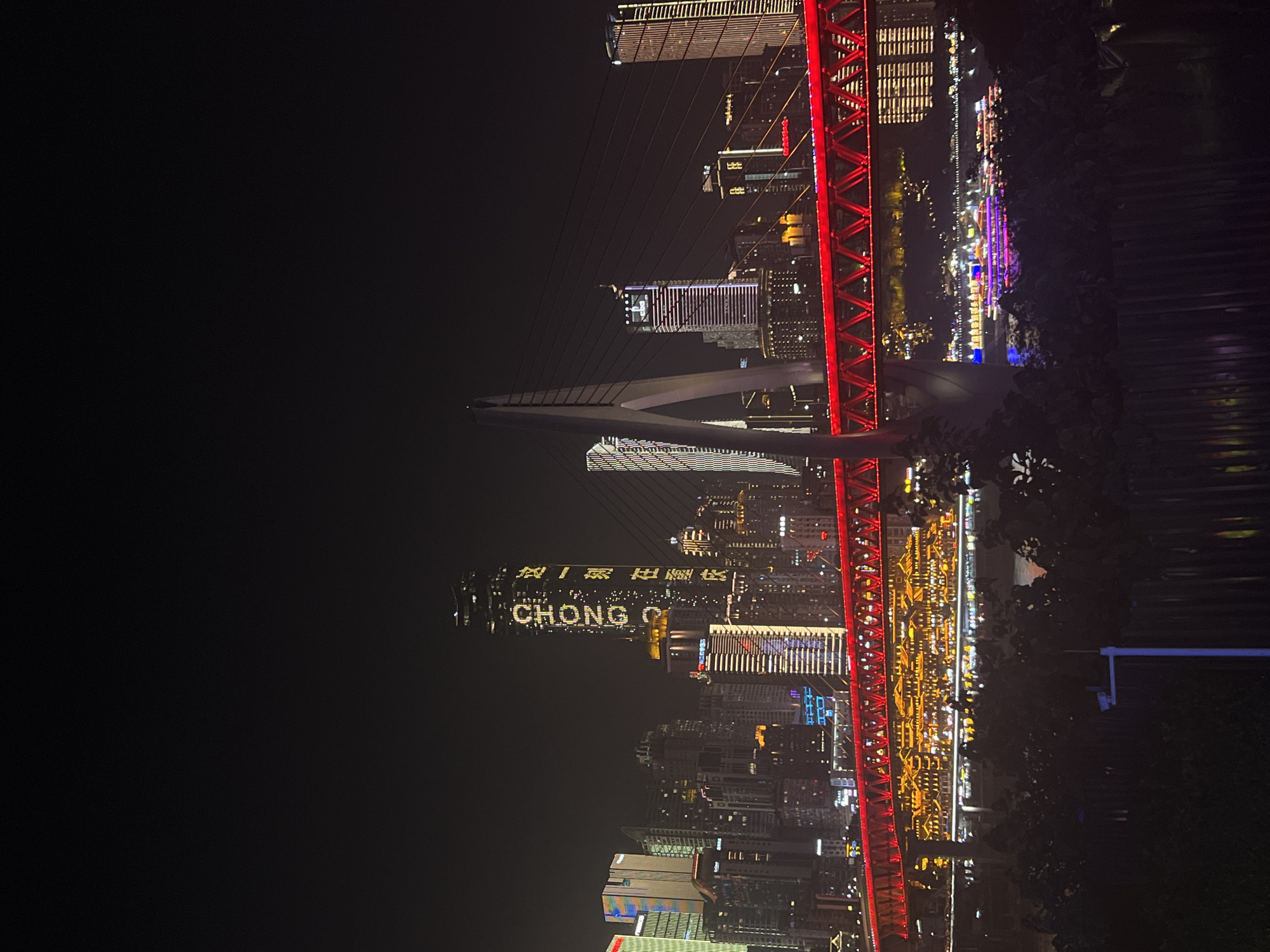 The image depicts a night scene in Jiangbei Cheng Street, China, showcasing a vibrant cityscape dominated by illuminated skyscrapers and a bridge. A tall skyscraper in the center features the word "CHONGQING" in large, bright yellow letters, with smaller Chinese characters above and below it. Adjacent to this, other buildings are adorned with colorful lights, creating a dazzling display. A prominent red suspension bridge stretches across the foreground, its structure outlined with bright red lights. Beneath the bridge, the river reflects the city's illumination, and several boats are visible on the water, some displaying colorful lights. The foreground is partially obscured by dark foliage and a black metal fence, suggesting the vantage point is from a dimly lit area overlooking the river. The overall atmosphere is one of a bustling, modern metropolis at night.