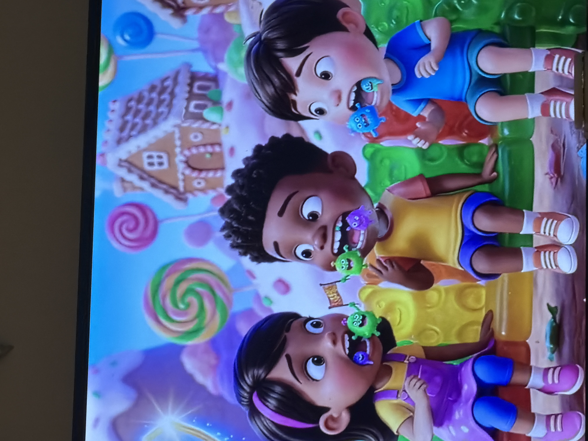 The image shows three cartoon children sitting on giant gummy bear-shaped benches in a whimsical candy land. The child on the left, a girl with dark hair and a purple dress over a yellow shirt and blue leggings, has a purple creature in her mouth. The child in the middle, a boy with dark curly hair wearing a yellow shirt and blue shorts, is holding a green creature with a flag in his mouth. The child on the right, a boy with dark hair in a blue t-shirt and shorts, has a blue creature and a green creature in his mouth. The background features a gingerbread house, giant lollipops, and candy-covered hills under a bright blue sky. There is no information in the image that provides the location in the city of Bac Nha Trang Ward, Vietnam.