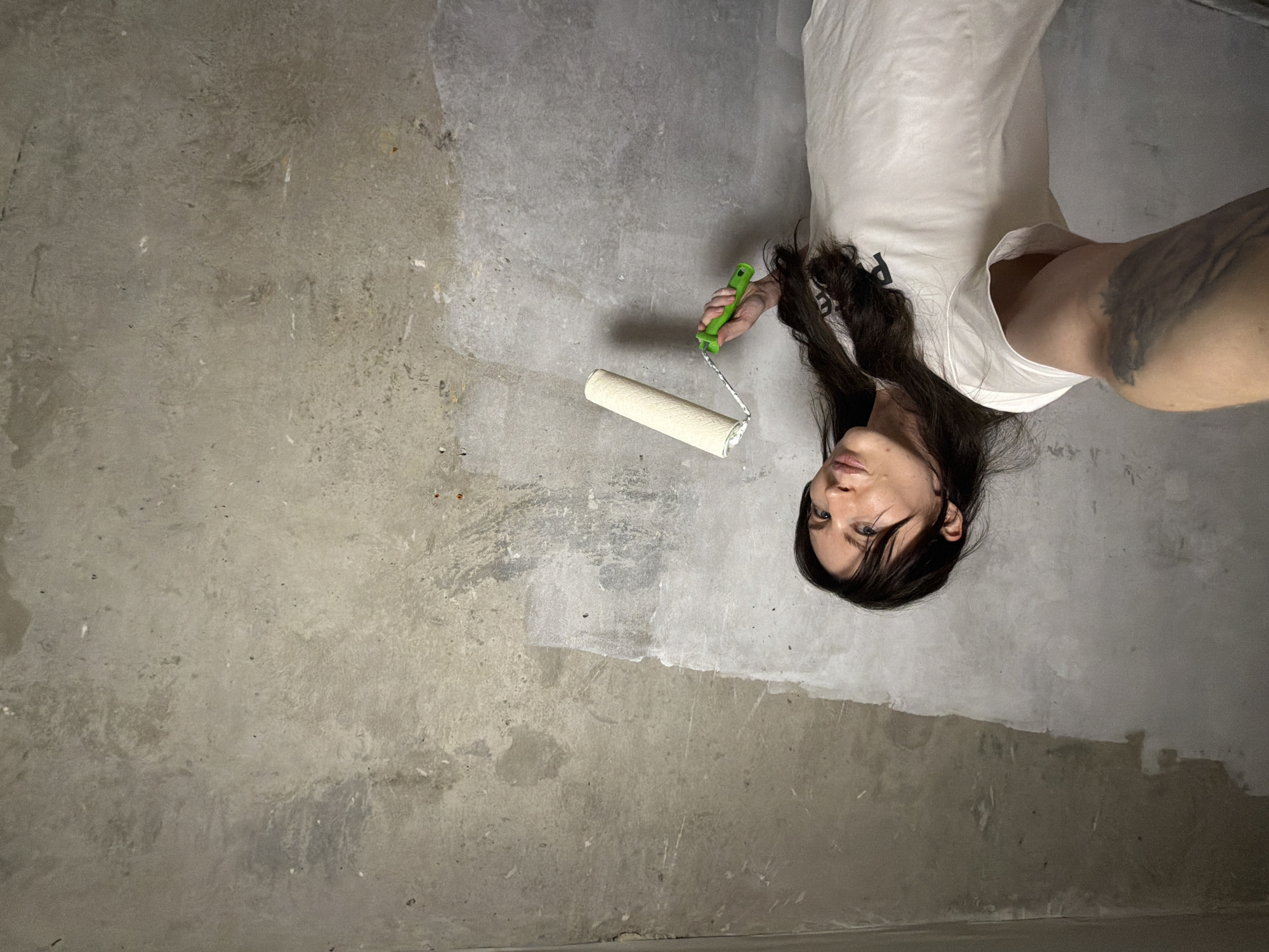 A female individual is captured in a selfie-style photograph within an indoor environment, likely a room undergoing renovation. The specific location within Perm, Russia, cannot be determined from the visual information provided. The subject, positioned on the left side of the frame, is a woman with long, dark hair, wearing a white short-sleeved t-shirt with dark lettering partially visible. Her right arm, extended towards the camera, reveals a dark tattoo on the inner forearm/bicep area. She holds a paint roller with a cream-colored roller head and a green handle in her left hand. The background consists of an unfinished, textured grey concrete or plaster wall. A significant portion of the wall, particularly from the lower left towards the center and extending upwards, shows evidence of being partially covered with a white substance, likely primer or paint, indicating ongoing surface preparation or painting work. The upper section of the wall and the ceiling line also appear to be unfinished. The individual is looking directly at the camera with a neutral expression.