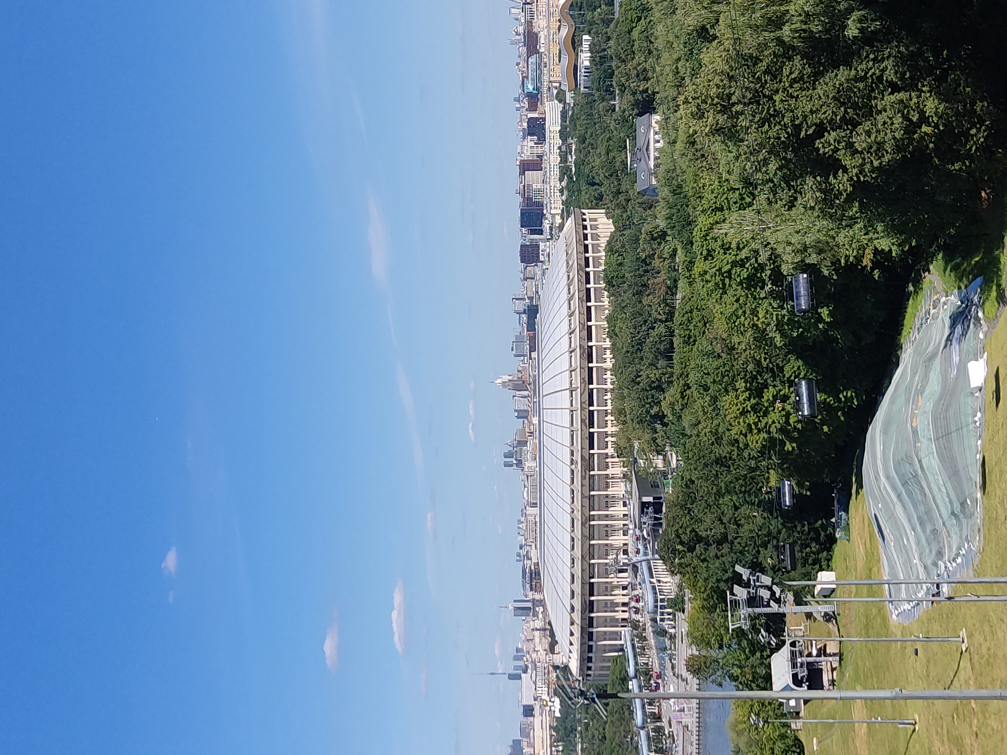 The image captures a panoramic view of Moscow, Russia, on a clear day. Dominating the foreground is the Luzhniki Stadium, a large, circular sports complex with a distinctive white roof supported by numerous columns. Surrounding the stadium is a lush green expanse of trees, indicating a park or natural area. In the mid-ground, a river flows, with a section of what appears to be a ski slope covered by a protective tarp in the lower left quadrant. Several ski lift cabins are visible suspended from cables, moving across the frame. Beyond the stadium and trees, the Moscow skyline unfolds, featuring a dense collection of modern buildings and skyscrapers, characteristic of a major urban center. The sky is a vibrant blue with a few scattered white clouds. The overall scene depicts a blend of urban infrastructure, natural landscapes, and recreational facilities within the city.