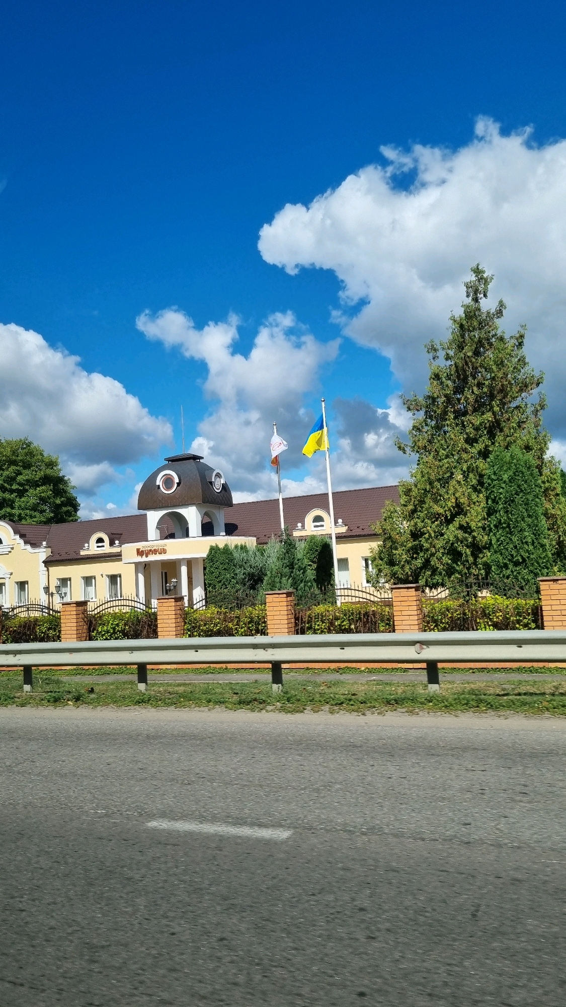 The image shows a yellow building with a brown roof and a dark dome on top. The building has several windows and arched entrances. Two flags are flying in front of the building, one is a white and red flag, and the other is the Ukrainian flag. A fence made of brick pillars and metal bars separates the building from the road in the foreground. A tall green tree stands to the right of the building. The sky is blue with scattered white clouds. The building appears to be located in Krupets, Ukraine, as indicated by the text on the building which reads "Крупець" (Krupets).