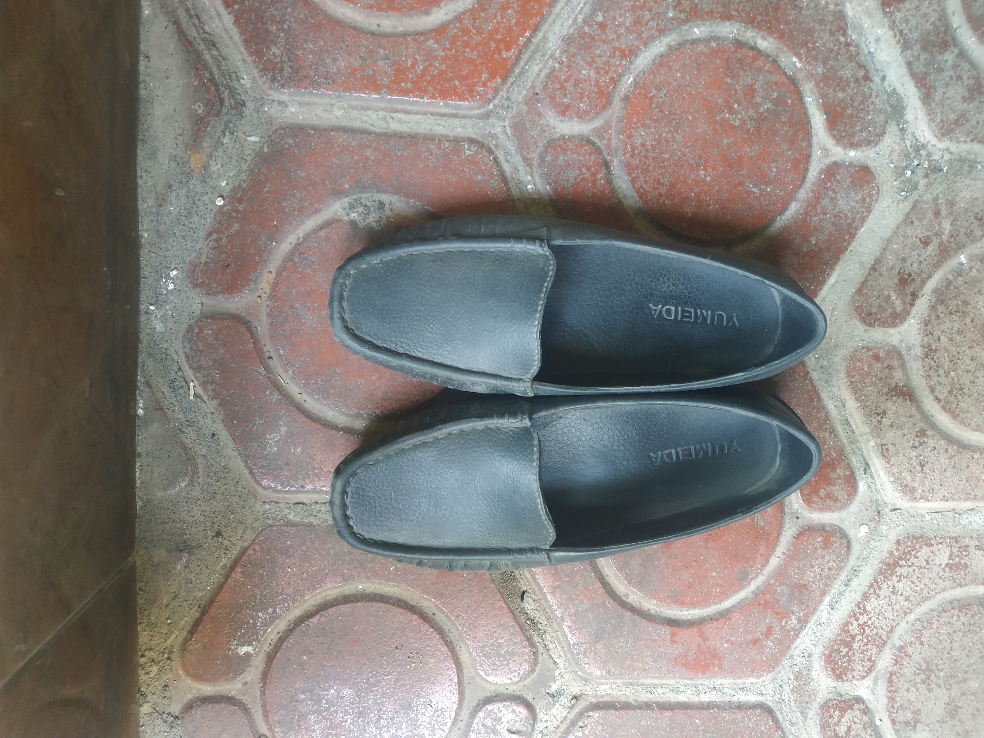 A pair of black slip-on shoes is shown from a top-down perspective on a red tiled floor. The shoes are positioned side-by-side, with the left shoe slightly above and to the left of the right shoe. The tiles have a hexagonal pattern with circular insets, and they appear somewhat worn and dusty. Above the tiled area, a darker, possibly concrete or wooden surface is visible, also showing signs of wear and dust. The lighting suggests an outdoor or semi-outdoor setting, likely during the day. The branding "YUMEIDA" is visible on the inner sole of both shoes. This scene could be located in a residential courtyard or pathway in Jember, Indonesia.