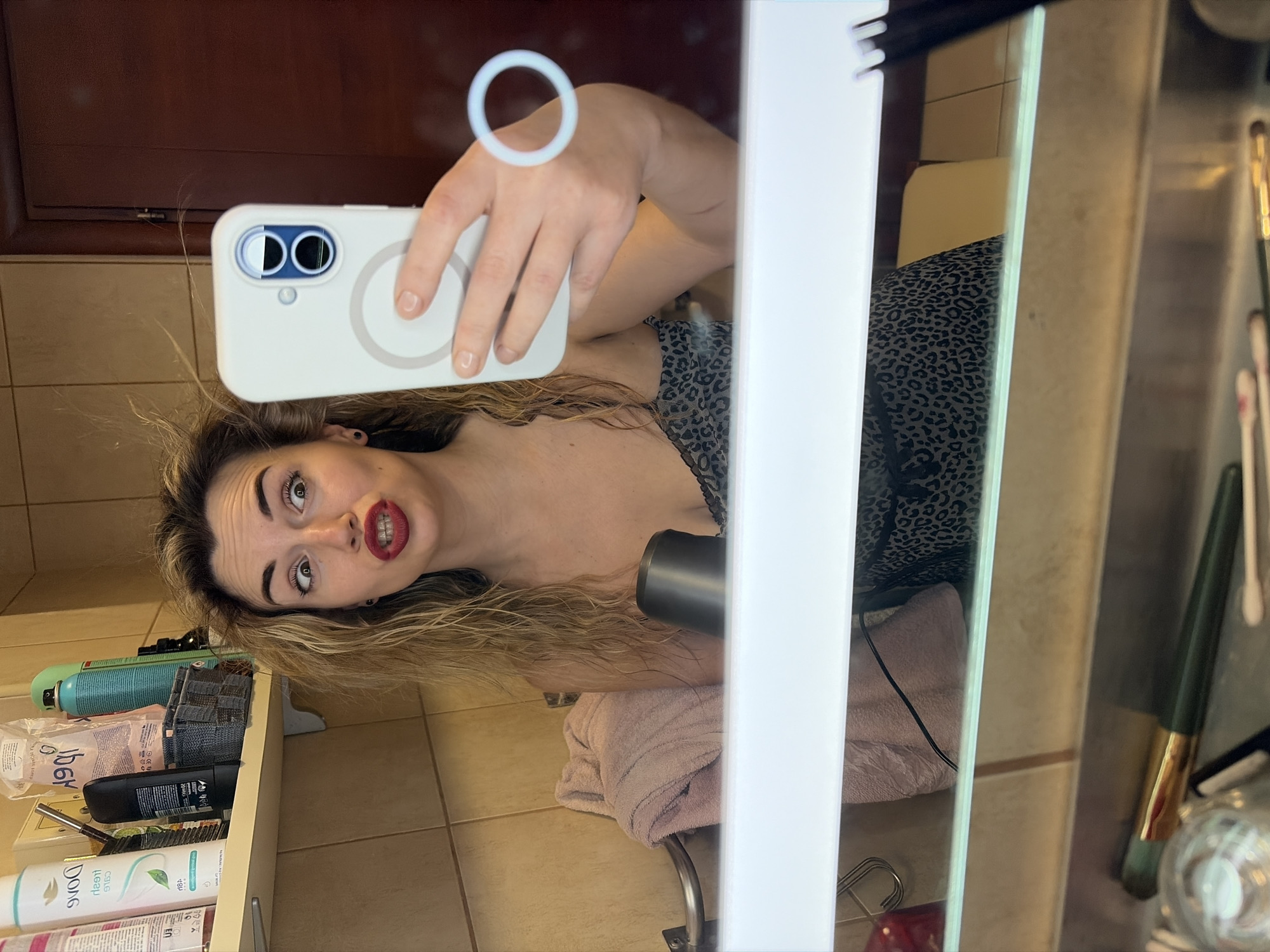 The image depicts a mirror selfie of a female in an indoor bathroom. The female has disheveled blonde, wavy hair and wears a black and gray leopard print top. Her face shows a playful expression with wide, crossed eyes and puckered lips, accentuated by dark red lipstick. She holds a white smartphone with a visible MagSafe ring in her right hand, capturing her reflection. Her left hand is visible holding a compact black hairdryer.
The reflected environment includes light brown square tiled walls and a dark brown wooden door. A white shelf is positioned above the mirror to the left, holding several personal care products, including a white Dove brand bottle and a blue aerosol can. A light pink towel is draped over a chrome towel bar below the shelf. An integrated light fixture within the mirror casts a bright horizontal bar across the frame. Below this bar, a vanity counter is visible, displaying a green and gold makeup brush and other small items like cotton swabs. The specific location in Budva, Montenegro, is not identifiable from the visual content of the image.
