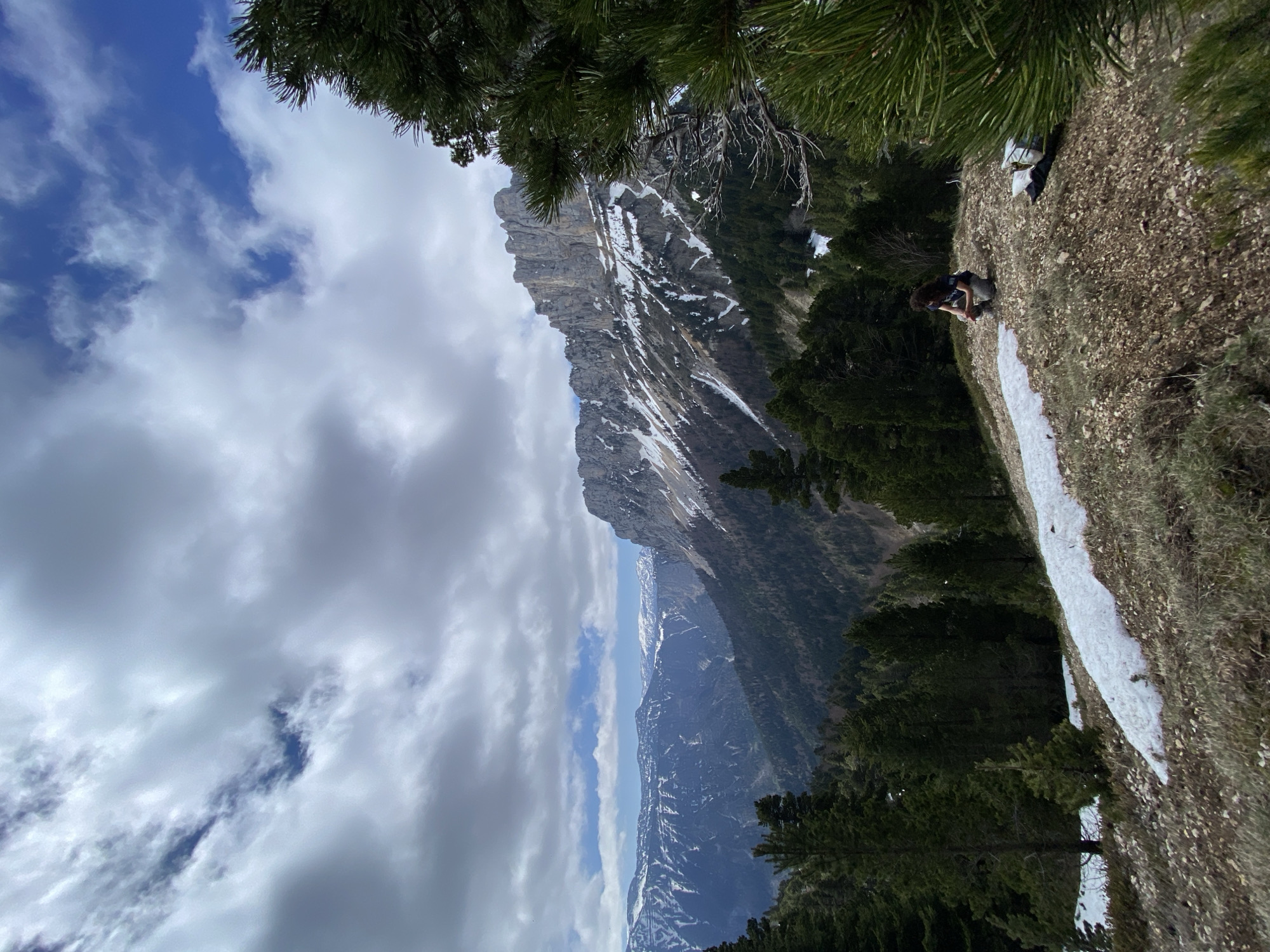 This outdoor photograph, taken in Chichilianne, France, captures a rugged mountainous landscape. The image is oriented such that the sky occupies the left side, while a steep, rocky slope with a person on it fills the right.
Dominating the central-left area, a towering, jagged mountain range rises vertically, its grey rock faces interspersed with significant patches of white snow. Dense green pine forests cover the lower slopes of these mountains. Further left, beyond the main peaks, distant mountain ranges are visible under a horizon line that appears midway on the left side of the frame. The sky, visible on the left, features a mix of bright blue patches and large, fluffy white and grey clouds, indicating partly cloudy conditions during daytime.
On the right side of the frame, closer to the viewer, the ground consists of dry, rocky earth with scattered bits of foliage. A distinct, narrow strip of snow lies on this slope, running roughly parallel to the bottom of the frame. A person is visible on this right slope, crouched low to the ground, wearing dark clothing. Next to them, a light-colored backpack is placed on the ground. Pine branches, with their dark green needles, frame the very top and bottom edges of the image, suggesting the viewpoint is from within a forested area looking out across the valley and mountains. There is no visible text. The scene conveys a sense of outdoor adventure and observation within a grand natural setting.