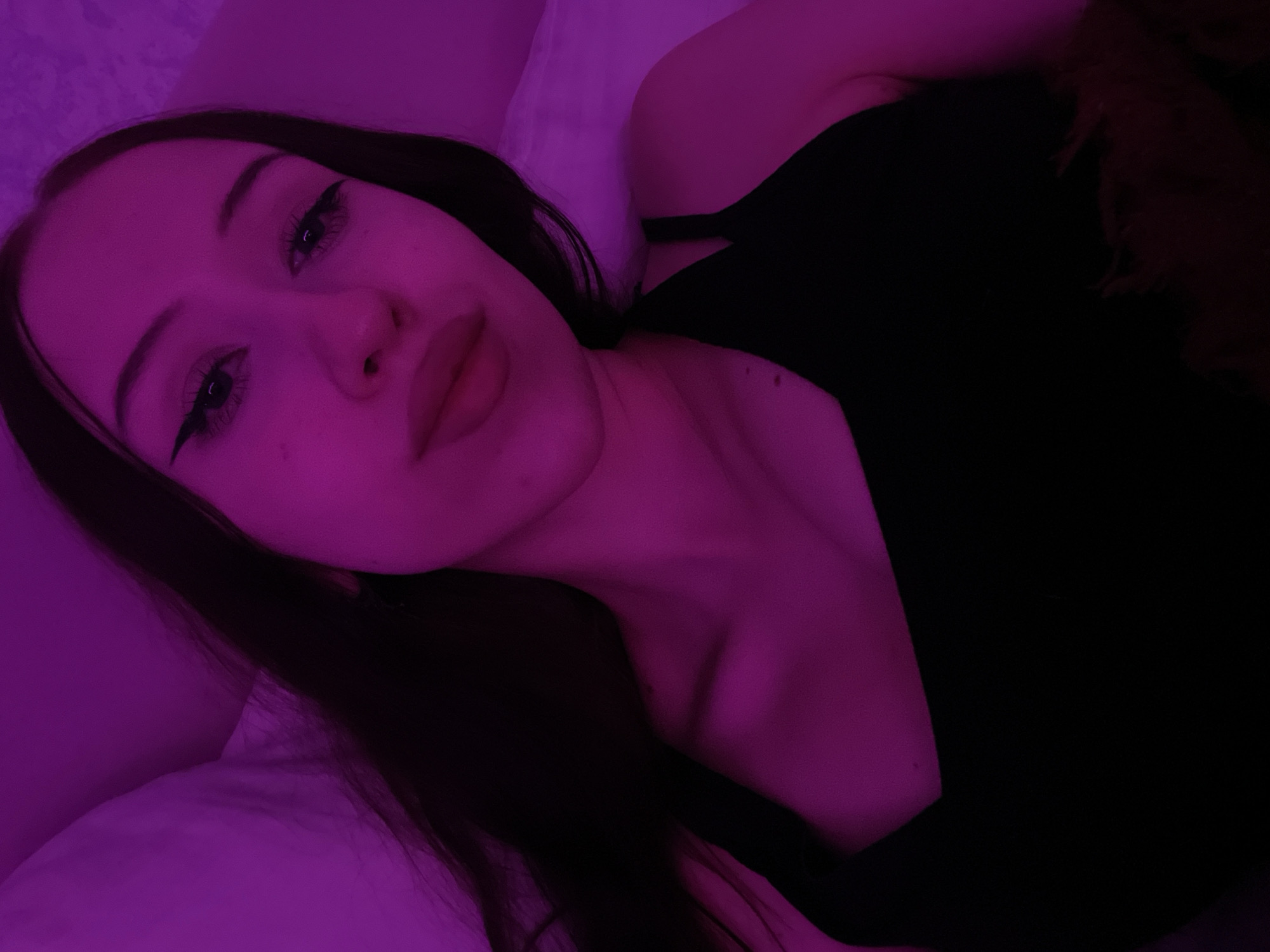 The image is a close-up, selfie-style portrait of a young woman, taken indoors. She has long, dark hair and is wearing a black top. Her makeup includes winged eyeliner and lip gloss. The lighting is predominantly pink, casting a purple hue over the scene. The background appears to be a bed with a white pillow. There is no indication of the city of Votkinsk, Russia, or any specific location within a city. The image focuses solely on the subject.