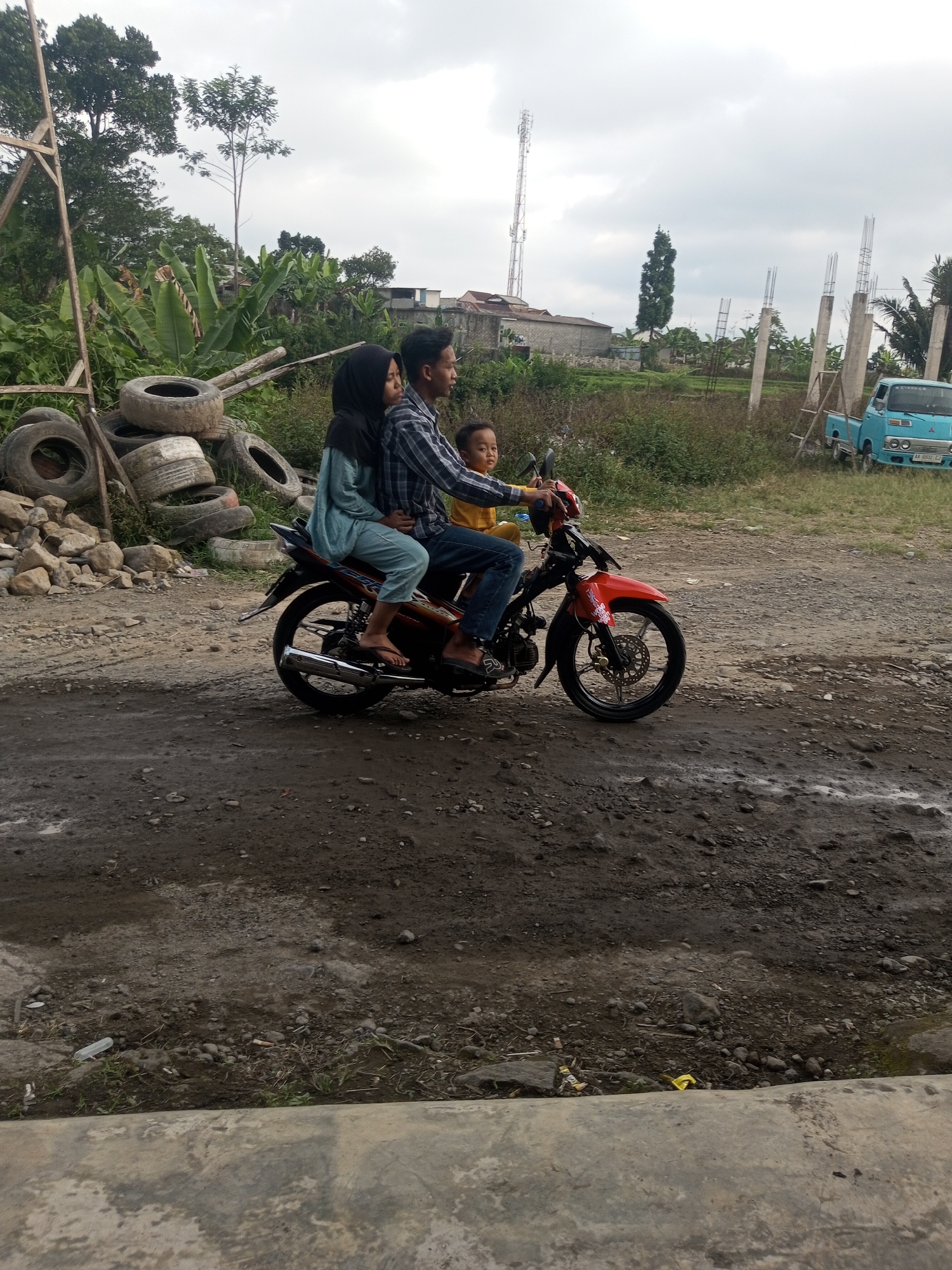 In Kalibeber, Indonesia, three individuals are riding a red motorcycle. A man in a checkered shirt is driving, with a woman wearing a hijab and a blue outfit behind him. A young child in a yellow shirt sits in front of the driver. They are on a dirt road with tires and debris piled to the left. In the background, there are banana trees, houses, a cell tower, and a light blue truck. The sky is overcast.