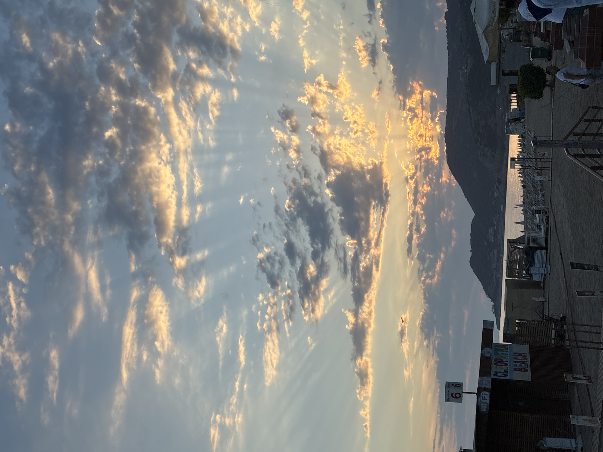 The image depicts a sunset scene at Cleopatra Beach in Alanya, Turkey. The sky is filled with dramatic clouds illuminated by the setting sun, casting golden and orange hues across the sky. Rays of light are visible piercing through the clouds. In the foreground, a beachside establishment is visible, featuring signage that reads "CLEOPATRA BEACH" and "WI FI". There are also numbers "66" on a sign, possibly indicating a beach section or facility number. The beach area appears to be set up with loungers and umbrellas, suggesting it is a popular tourist destination. A dark silhouette of a mountain range borders the horizon on the right side of the image. The overall atmosphere is serene and picturesque, characteristic of a peaceful evening at the beach.