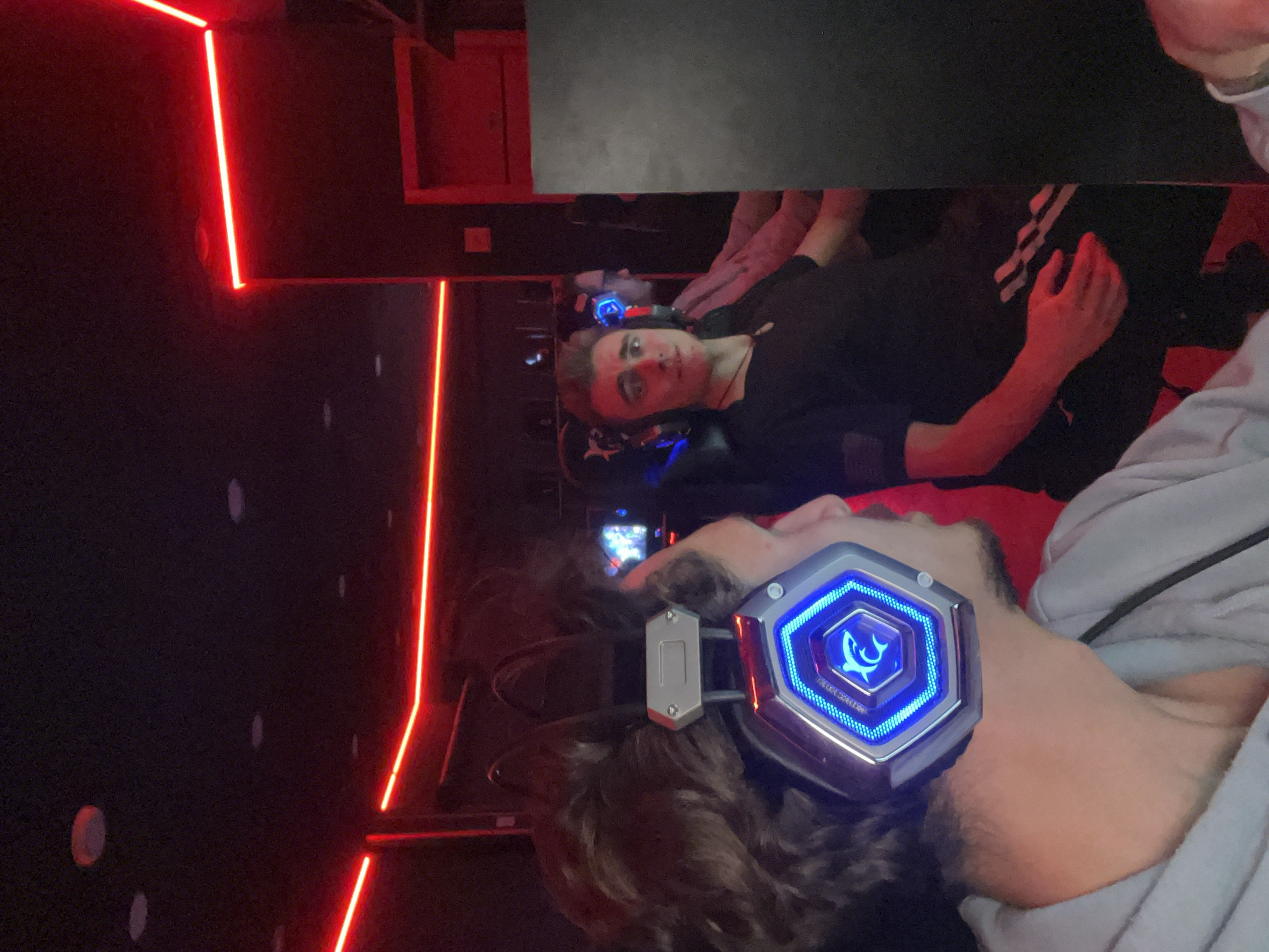 Two young men are in what appears to be a gaming room, illuminated by red neon lights. One man is wearing headphones with blue LED accents. The image might be taken indoors, possibly in Budva, Montenegro. There are reflections in a dark surface overhead.