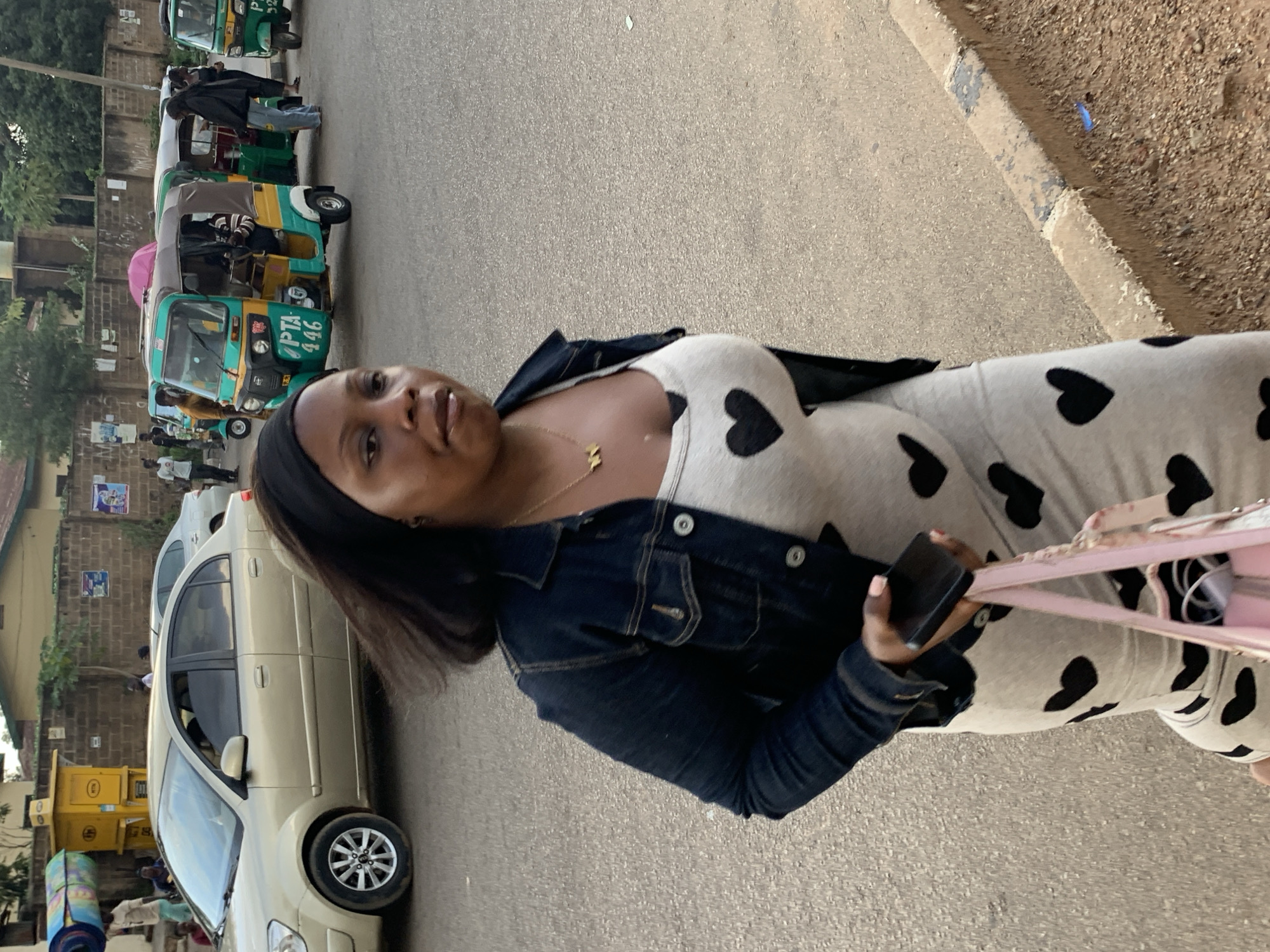 A medium shot captures a Black woman walking on a paved street in Jos, Nigeria. She is wearing a black denim jacket over a light-colored dress adorned with black heart patterns. She has a black headband and a gold necklace with an "M" charm. In her right hand, she holds a dark smartphone, and in her left, a light pink bag. The street is lined with a concrete curb and gravel. In the background, a beige sedan is parked, and several open-air auto-rickshaws, known locally as "Keke Napep," are visible, with passengers inside some. Buildings and a yellow kiosk are also present. The overall scene depicts a typical street view in Jos.