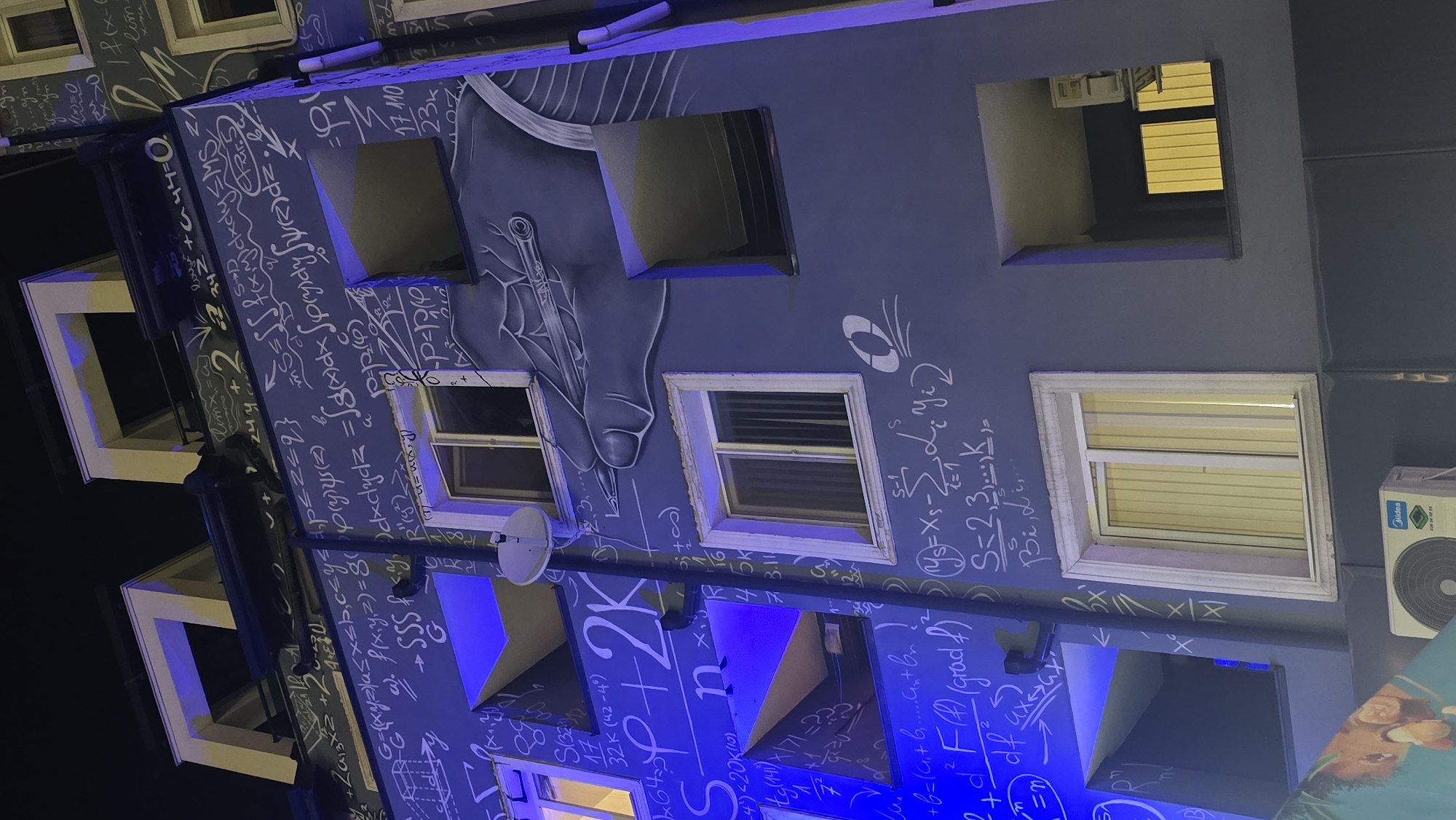 The image shows a building in Sofia, Bulgaria, decorated with mathematical equations, formulas, and illustrations. The building is several stories high, with windows visible on each level. The walls are painted blue and adorned with white mathematical expressions. A drawing of a hand holding a pen is also visible on the building's facade. An air conditioning unit is installed on the side of the building. The photo appears to be taken at night or in low light conditions.
