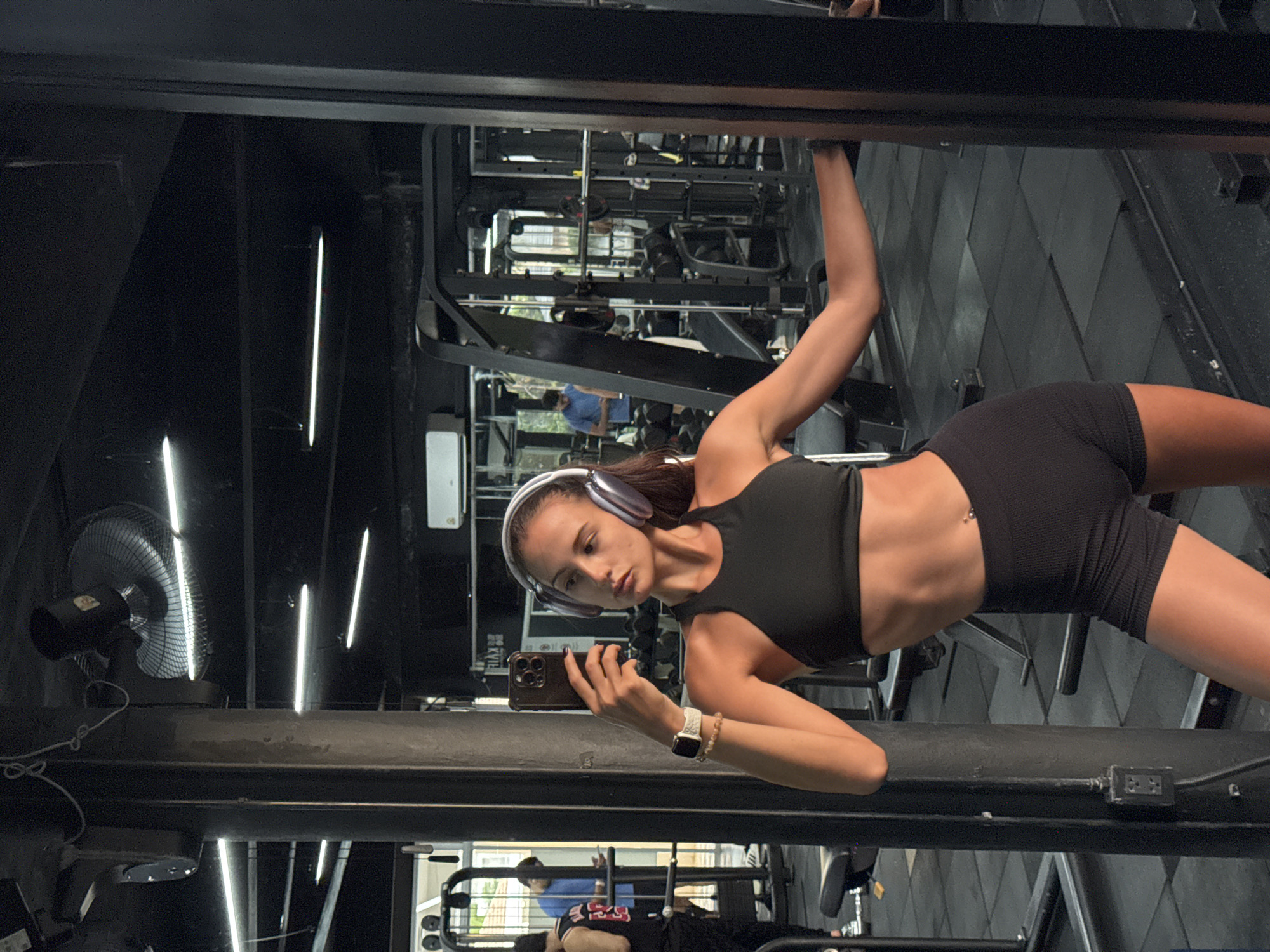 A young woman in a black workout top and shorts is taking a selfie in a gym, standing next to gym equipment. She is wearing headphones and a watch. The gym has various weight machines, a fan, and fluorescent lights. The reflection in the mirror shows the gym's interior and possibly other gym-goers. The location is Kathu, Thailand.