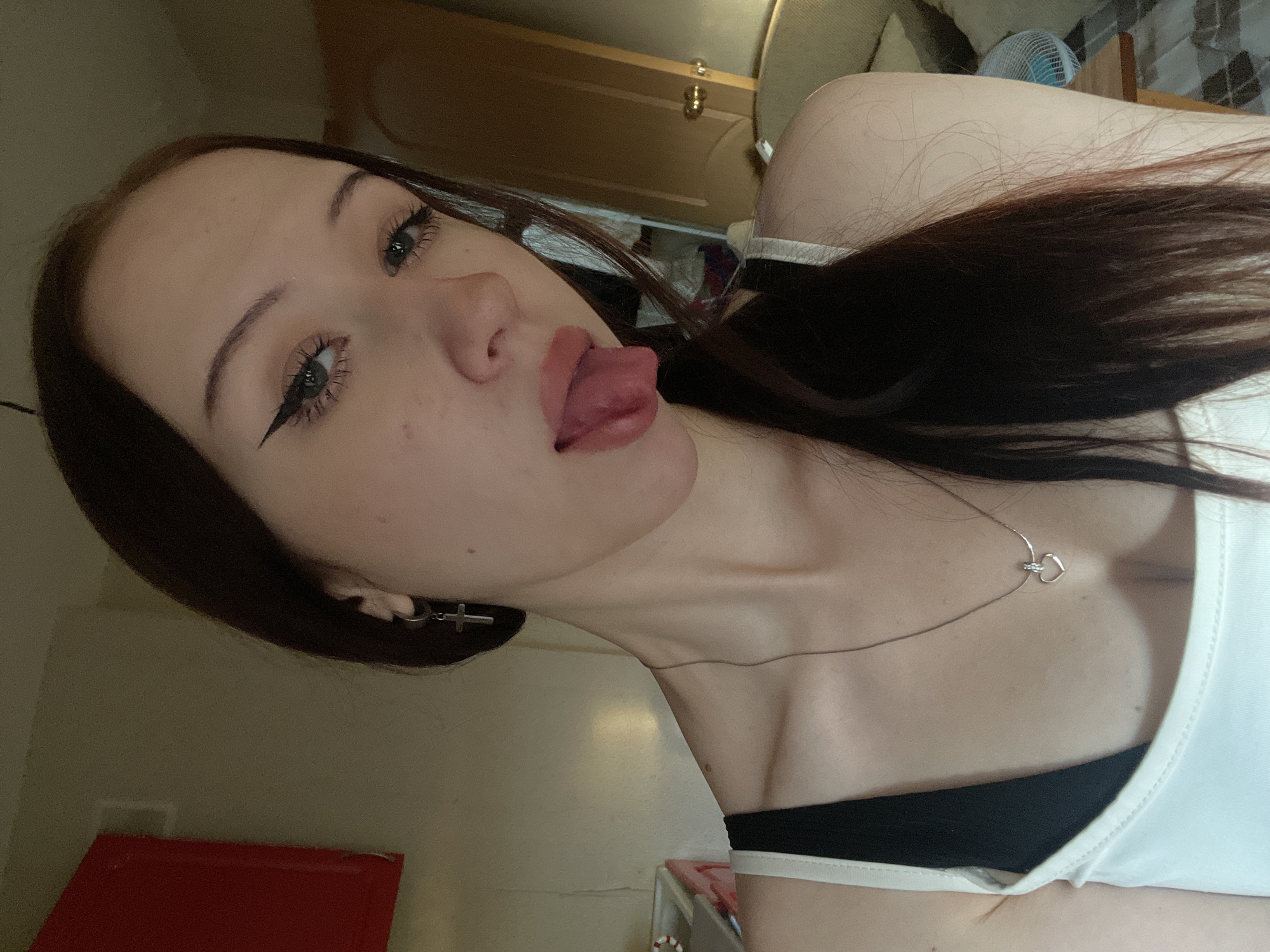 The image is a close-up selfie of a young woman indoors, likely in a residential setting. She has long, dark brown hair, styled with a side part. She is wearing a white tank top over a black bra and a silver necklace with a heart pendant. She has on winged eyeliner and her tongue is sticking out slightly. A silver cross earring is visible on her left ear. The background appears to be a room with a wall, a closed door, and a glimpse of furniture. A red object, possibly a cabinet or a picture frame, is mounted on the wall to the left. The lighting is dim and appears to be natural or from an artificial indoor source. There is no specific indication of the city of Votkinsk, Russia within the image itself.