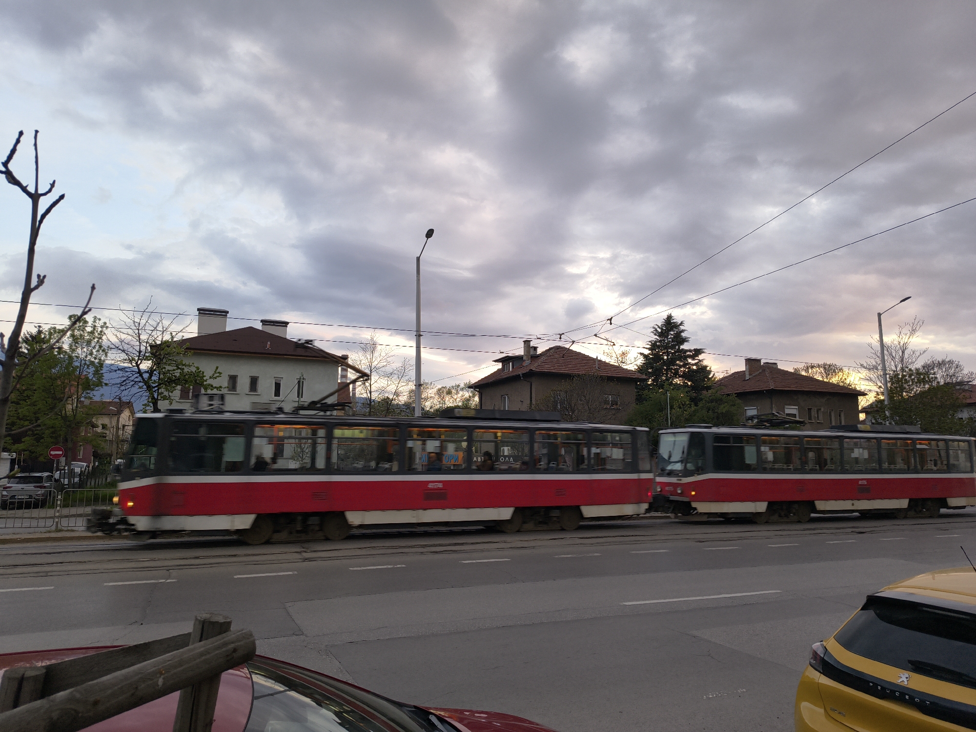 The image depicts an urban street scene in Sofia, Bulgaria, likely at dusk, characterized by an overcast sky with subtle warm hues on the horizon. The central focus is on two red and white trams, a prominent feature of the city's public transport, moving on parallel tracks. The tram in the foreground is slightly blurred, indicating motion, with its interior lights illuminating passengers within. Visible through its windows are partial Cyrillic texts "ИСТОРИ" and "АВТО ОЛА," possibly parts of advertisements or route details. The tram behind it is numbered "4075" on its front and also has its lights on.
The street in the foreground features a two-lane road with white dashed markings. Parts of two cars are visible: a yellow Peugeot 208 on the right, with "PEUGEOT" clearly visible on its tailgate, and a red car in the bottom left corner. To the far left, behind a metal fence, a silver Hyundai car is parked, accompanied by a red circular 'no entry' sign with a white horizontal bar.
In the background, residential buildings with distinct dark red-tiled roofs and multiple chimneys are visible, nestled amidst various trees, some bare and others with green foliage. Lampposts and overhead electrical wires, typical for tram systems, crisscross the cloudy sky. The scene captures a moment of active public transit in a residential part of the city.