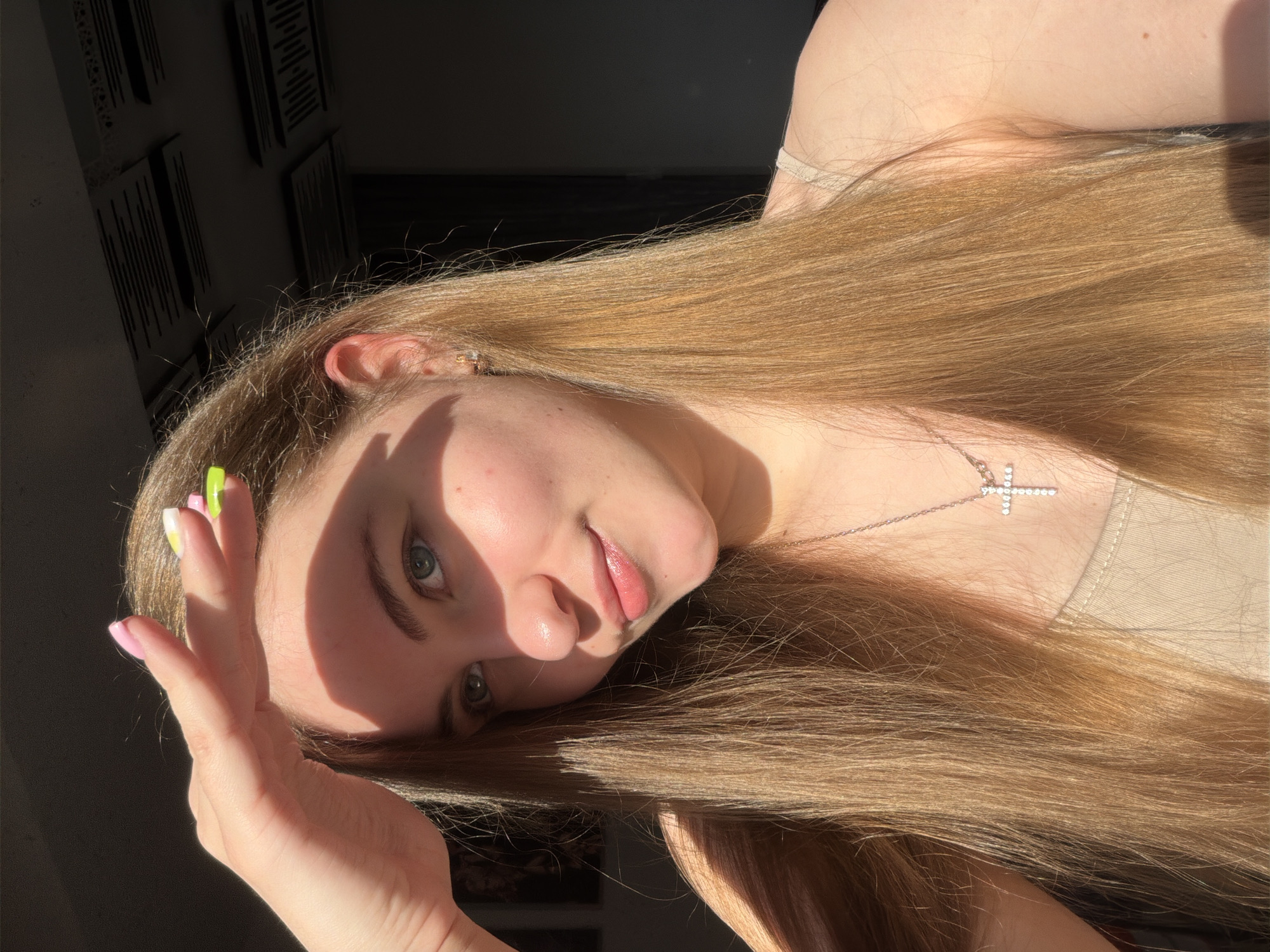 A young woman with long, light brown hair and blue eyes is captured in a close-up selfie. She is shielding her face from the sun with her hand, which has brightly colored nails. She is wearing a beige tank top and a diamond-encrusted cross pendant. The background appears to be indoors with some acoustic panels on the ceiling, suggesting a home or studio environment. There are no discernible landmarks or elements that would indicate a specific location within the city of Dnipro, Ukraine. The lighting suggests it is daytime, with sunlight casting shadows on her face.