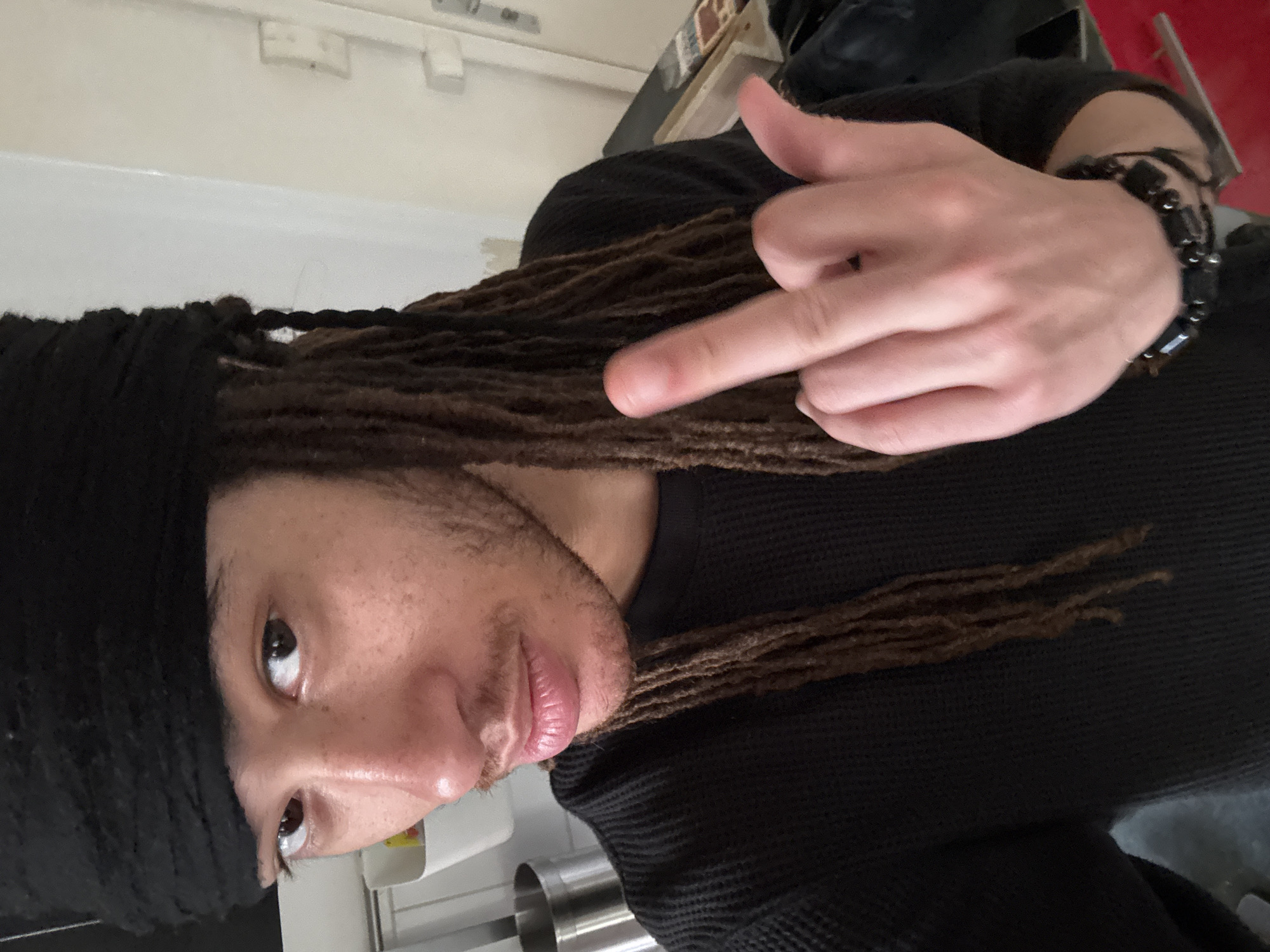 A person with dark hair styled in dreadlocks and wearing a black head wrap and a black long-sleeved shirt is gesturing with their hand, appearing to extend the middle finger. The person is also wearing a black bracelet on their wrist. The background is out of focus but appears to be an indoor space with white walls and some visible objects. The location is not determinable as Paris, France.