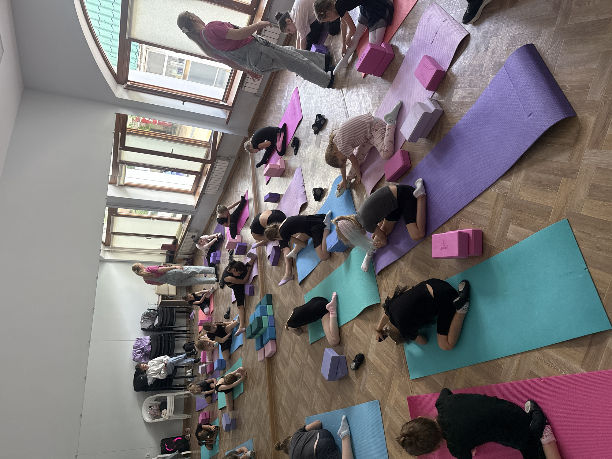 A dance class is in session in a room with a wooden floor and a large window. The instructor, a woman with a pink shirt and overalls, is standing at the front of the class. There are approximately 15 students, mostly young girls, on yoga mats in rows. Each student has blocks near them. The students are stretching. A large mirror can be seen on the left side of the frame.