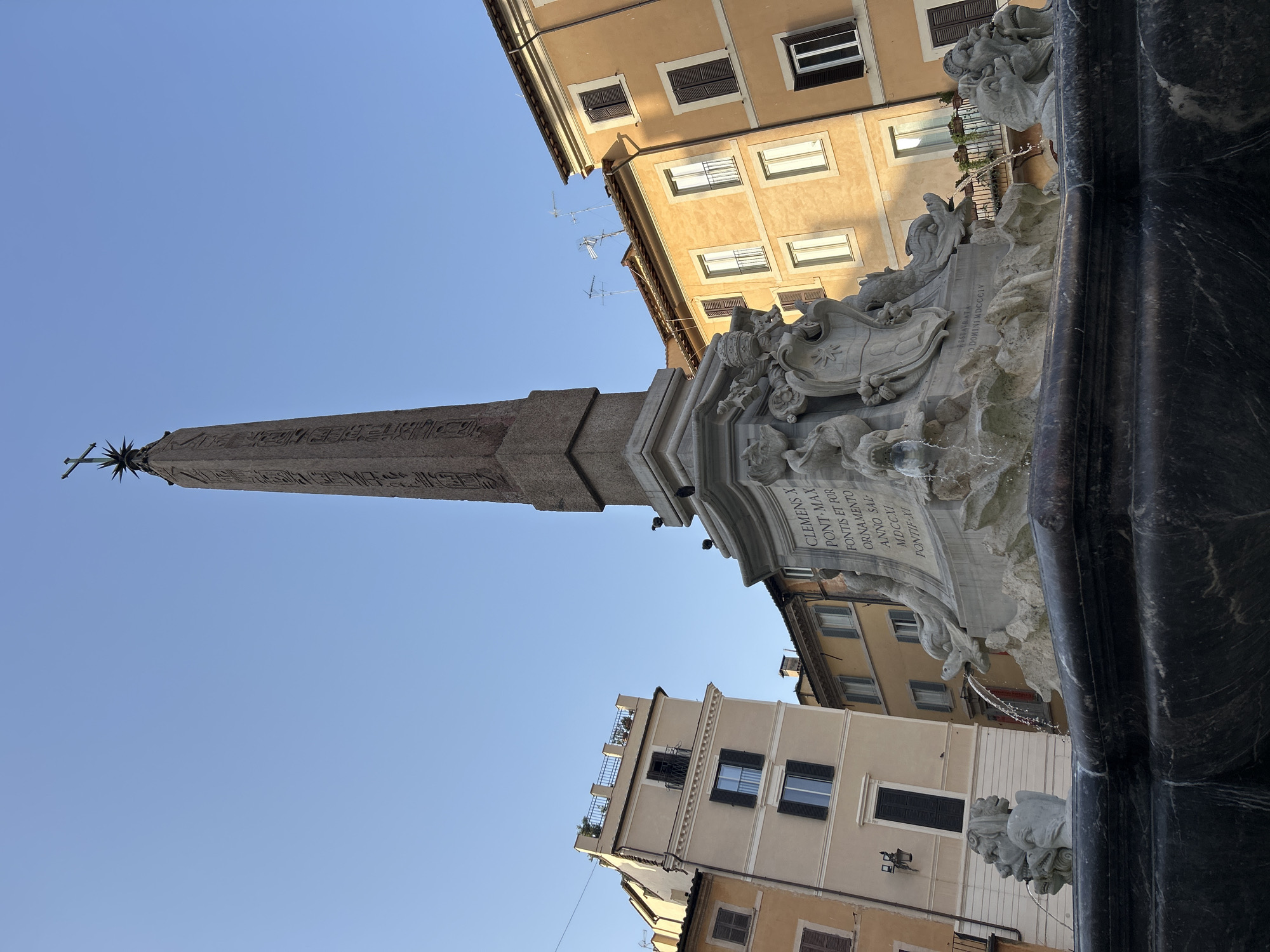 The image depicts the Fontana dei Quattro Fiumi (Fountain of the Four Rivers) in Piazza Navona, Rome, Italy. The fountain, designed by Gian Lorenzo Bernini, features a large granite obelisk at its center, topped with a cross and a star. The base of the obelisk is a complex arrangement of sculpted figures representing the four major rivers of the continents known at the time: the Nile, the Ganges, the Danube, and the Río de la Plata. Water cascades from the mouths of mythical creatures and the sculpted figures into the fountain's basin. Surrounding the fountain are ochre and beige buildings with shutters and balconies, indicative of Roman architecture. The sky is clear and blue. Several pigeons are perched on the fountain's structure. The image captures a daytime scene, with ample sunlight illuminating the architecture and the water feature.