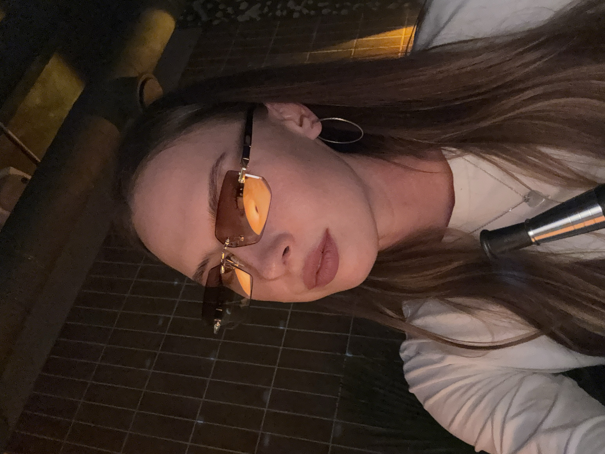A woman is shown from the chest up, wearing sunglasses and a white shirt. She has long brown hair and is wearing hoop earrings and a necklace with a heart-shaped pendant. She appears to be holding a dark cylindrical object. The background consists of a dark, tiled wall and some illuminated structures. The scene suggests an indoor setting, possibly a bar or lounge, with artificial lighting. There is no specific information to suggest the location is the city of Perm, Russia.