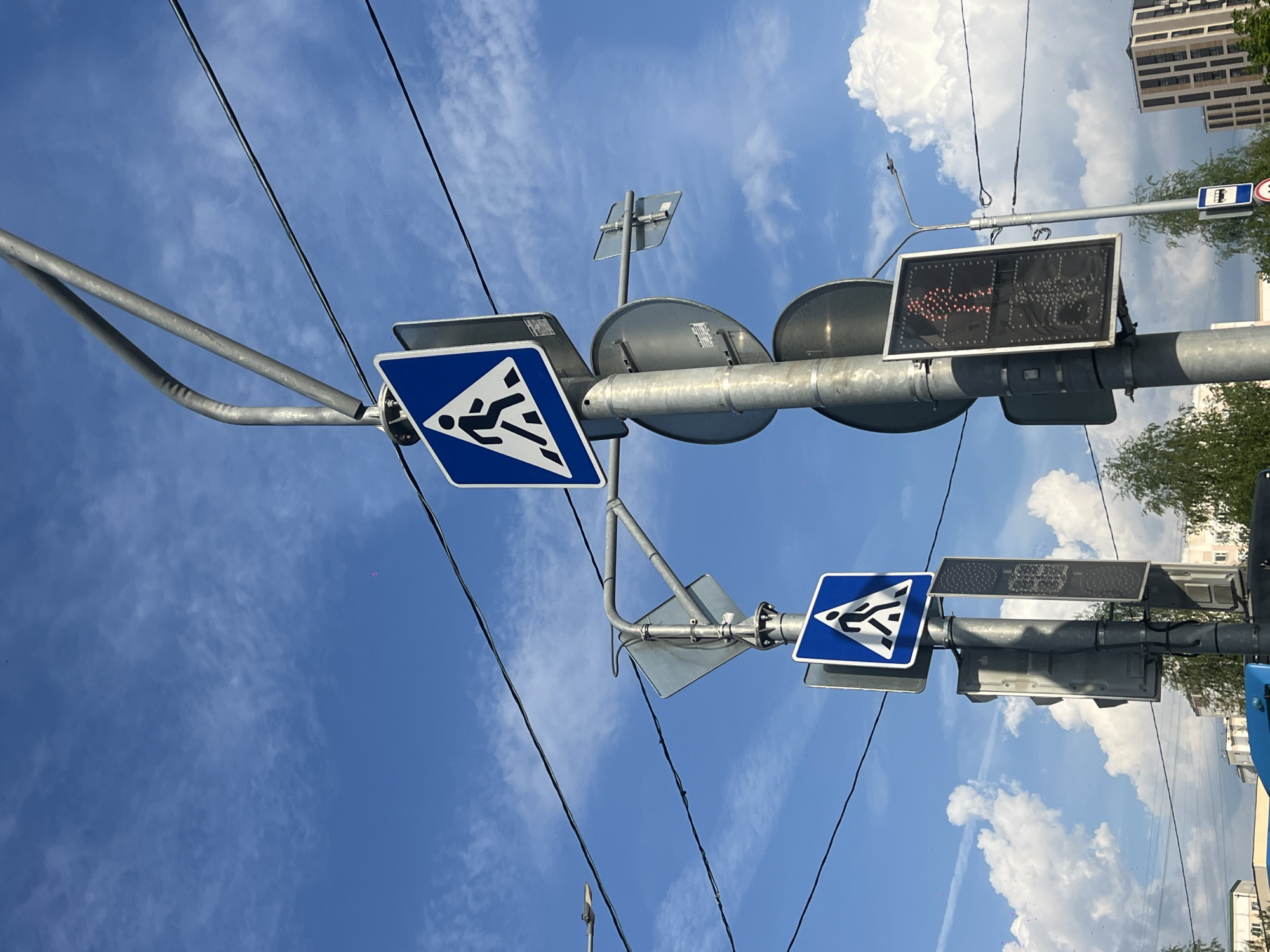 The image shows a pedestrian crossing sign in Moscow, Russia. The sign features a blue square with a white triangle inside, depicting a figure running. There are two such signs mounted on a metal pole, one on each side. The pole also holds a pedestrian traffic light with a display showing a walking figure illuminated in red dots. Overhead electrical wires are visible against a blue sky with scattered clouds. In the background, buildings and trees are partially visible.