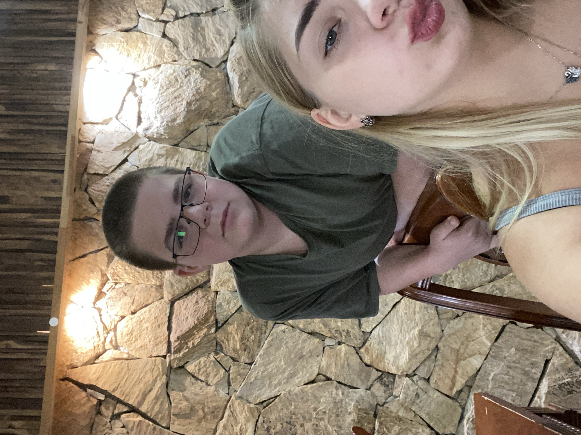In an indoor setting with a stone-tiled wall and a wooden ceiling, two individuals are visible. A young person with short hair and glasses is positioned in the background, leaning on a wooden chair. In the foreground, a young person with blonde hair is in focus, making a kissing gesture. The overall lighting suggests an evening or indoor ambiance. The location is identified as Sredets, Bulgaria.
