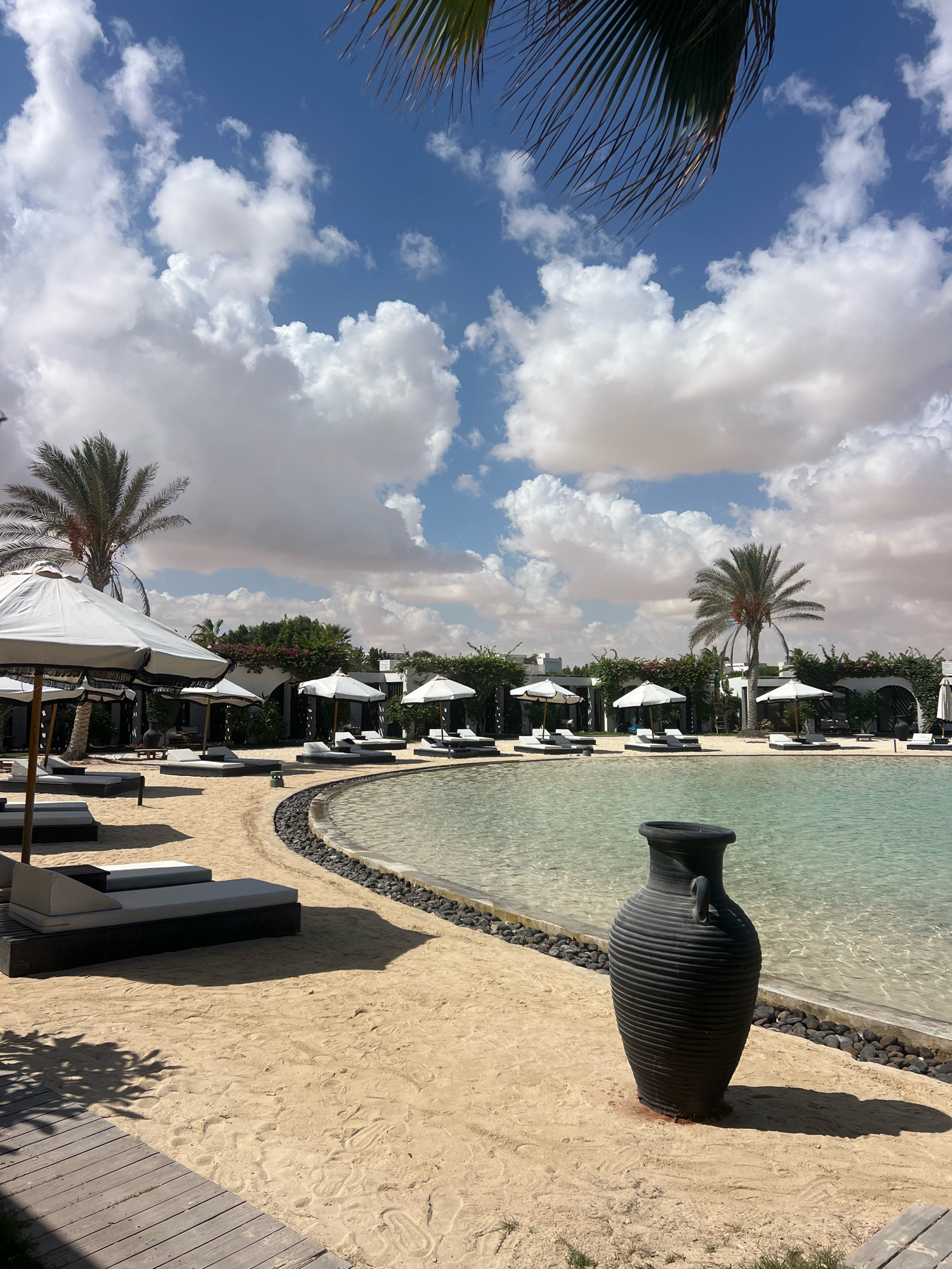 A serene outdoor scene is depicted in the city of Sidi Abdel Rahman, Egypt, under a bright blue sky dotted with fluffy white clouds. In the foreground, a light wooden walkway leads to a sandy beach area. Several lounge chairs with gray cushions are arranged on the sand, some with white umbrellas providing shade.
A curved pool with clear turquoise water is bordered by dark stones. The water's surface reflects the light and hints at the sandy bottom beneath. A large, dark-colored, ribbed vase stands on the sand near the edge of the pool, casting a distinct shadow.
In the background, more lounge chairs and umbrellas are visible, along with the facade of buildings and lush greenery, including palm trees. The overall atmosphere is one of a tranquil resort or beach club setting, inviting relaxation and enjoyment. There are no people visible in the image.