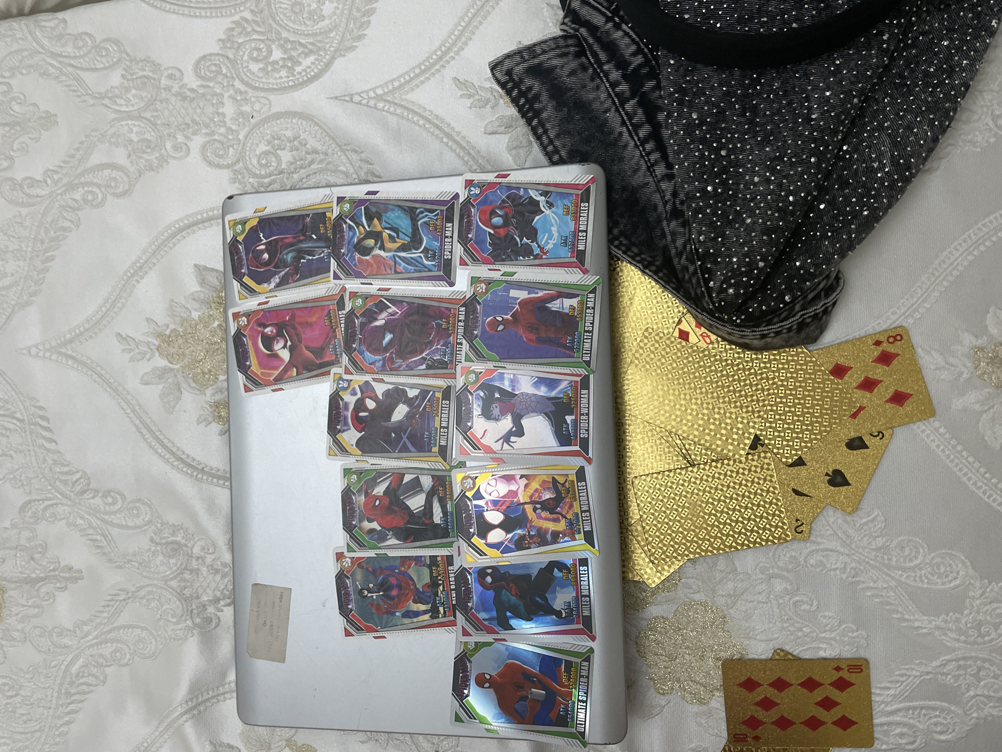 An indoor, top-down shot taken in Ihtiman, Bulgaria, displays a collection of items arranged on a white bedspread with an ornate, light gray and gold patterned design. Dominating the center is a silver laptop or tablet, serving as a base for approximately fifteen Spider-Man themed trading cards. These colorful cards feature various iterations of Spider-Man characters, including Miles Morales, Spider-Woman, Ultimate Spider-Man, and a character identified as "NENI DARKER". Each card displays "ATK" (Attack) and "DEF" (Defense) statistics, with values ranging from 248,000 to 899,000.
To the upper right, a section of a dark gray or black denim garment, heavily embellished with numerous small, sparkling rhinestones or studs, is visible. Adjacent to this garment and extending onto the bedspread are several gold-colored playing cards. Some cards are face up, revealing suits and numbers such as the 2 of Spades, 5 of Spades, 8 of Diamonds, and 10 of Diamonds. The gold cards have an intricate geometric pattern on their backs. A small, faded sticker with partially legible text, including "sdny" and "https://...", is affixed to the lower left corner of the laptop. The overall scene suggests a casual arrangement of personal belongings.