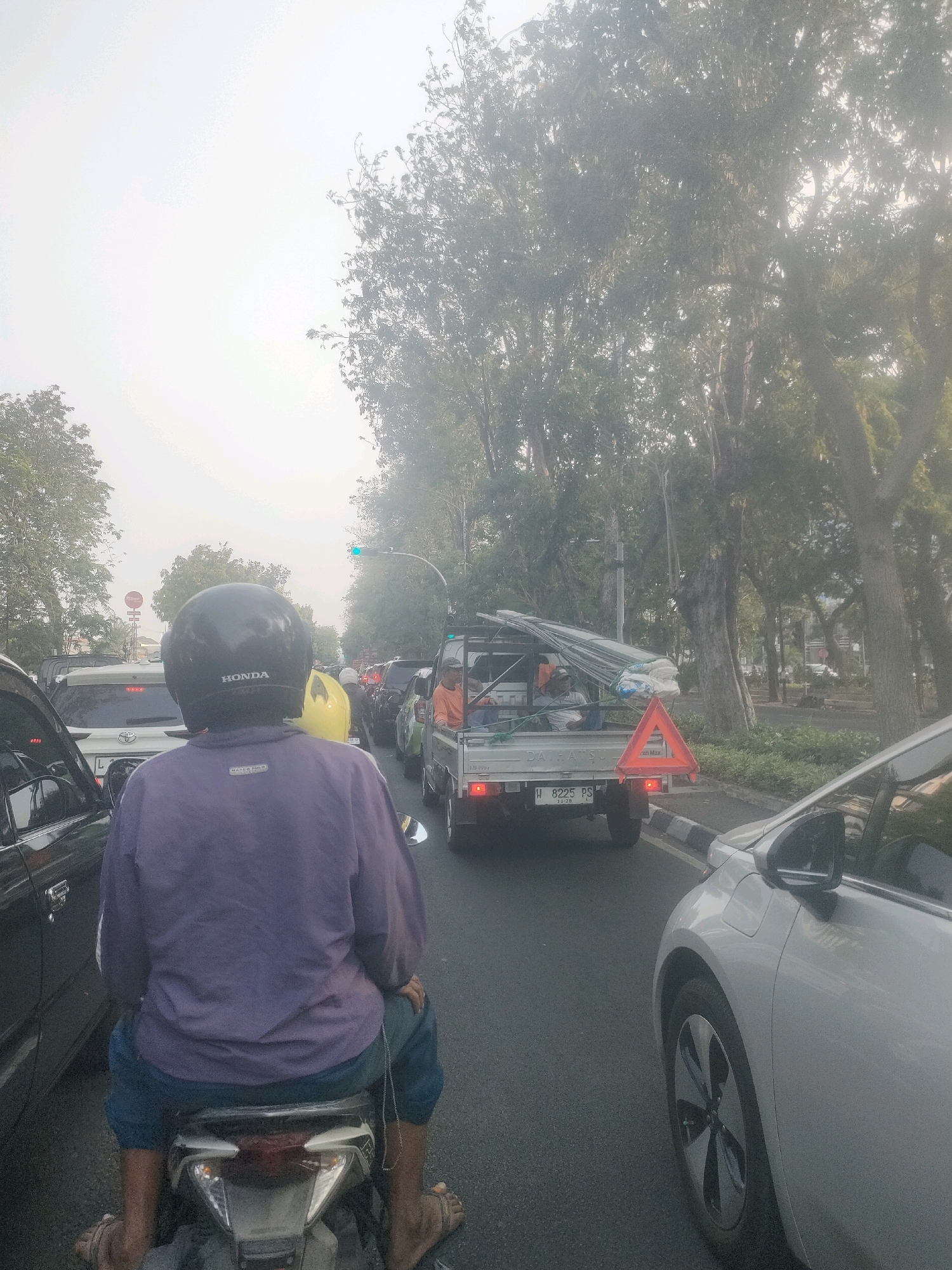 This image depicts a traffic jam on a street in Surabaya, Indonesia, likely on a hazy or smoggy day. In the foreground, a person wearing a purple long-sleeved shirt and blue pants is seated on a motorcycle, facing away from the camera. They are wearing a black helmet with "HONDA" written on it. Beside the motorcycle, to the left, is the side of a dark-colored car. Ahead of the motorcycle, several other vehicles are visible, including a white Toyota SUV and a light-colored pickup truck loaded with metal pipes and construction materials, identified by a license plate reading "W 8225 PS". A red triangular reflector is attached to the rear of the pickup truck. Trees line the right side of the road, and a traffic light, showing green, is visible in the distance on the right. The overall atmosphere suggests slow-moving traffic within an urban environment.