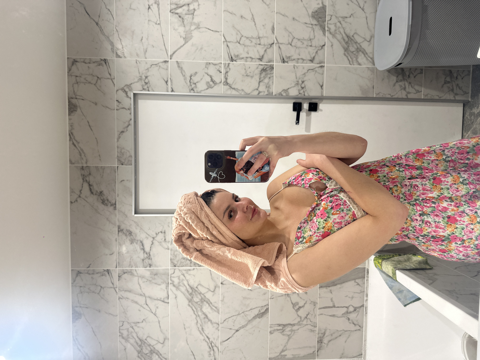 A young woman is taking a selfie in a bathroom. She is wearing a floral-print slip dress and a peach-colored towel is wrapped around her head. She is holding a smartphone in her right hand, which is angled to capture her reflection in the mirror. The bathroom features marble-patterned tiles on the walls and a white bathtub. A white door with black handles is visible behind her, and a grey laundry hamper is to the right of the door.