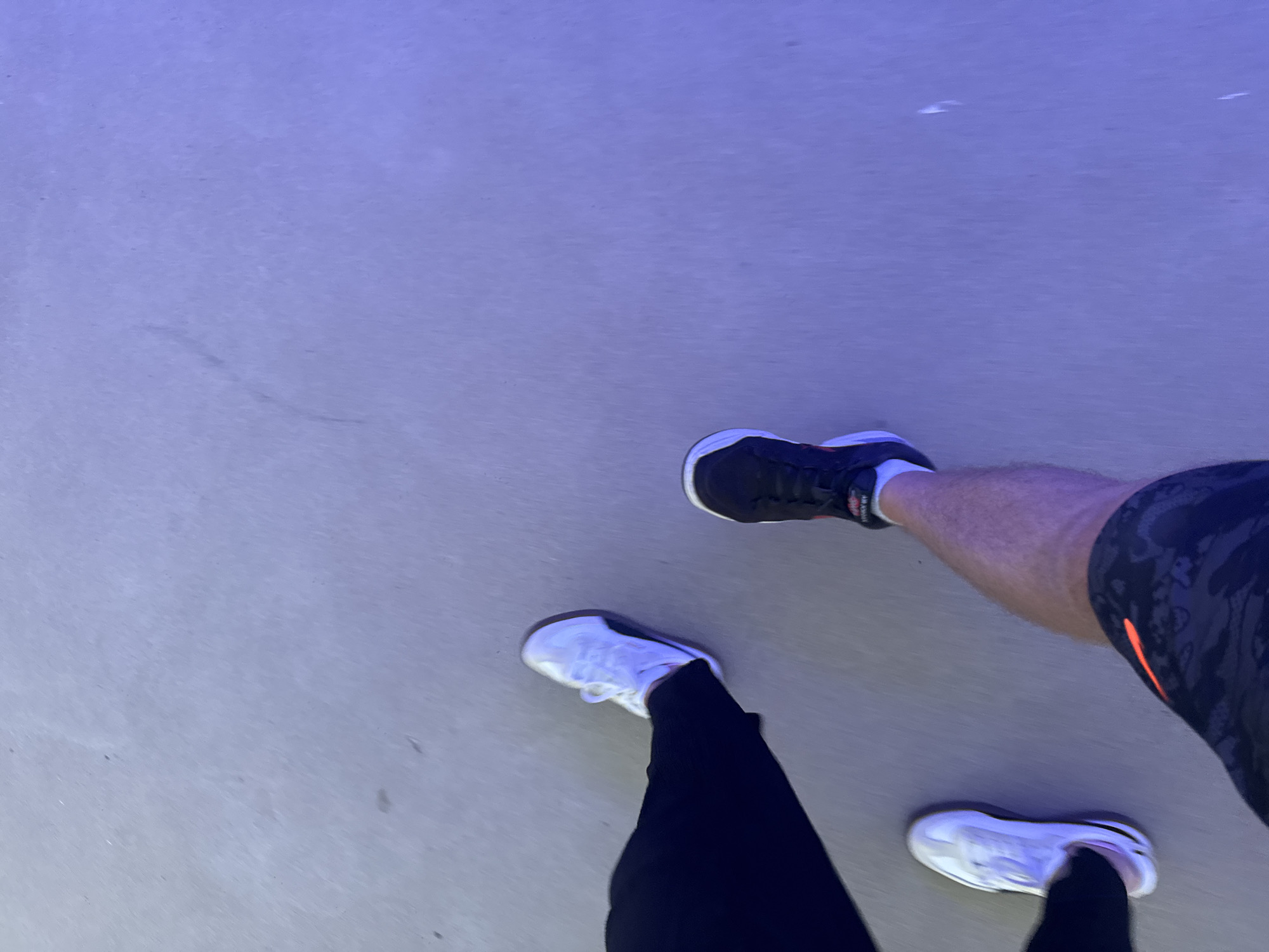 Two pairs of feet are visible, standing on a light-colored, smooth surface, possibly asphalt or concrete, illuminated by a blueish light. On the left, a person's left foot is shod in a white sneaker with black accents. The person is wearing dark-colored trousers. To the right and slightly behind, another person's right foot is visible, wearing a black and white sneaker with red accents. This person is wearing patterned shorts. The overall scene appears to be outdoors, with the ground reflecting the ambient blue light. The specific location in Tallinn, Estonia cannot be determined from the provided image.
