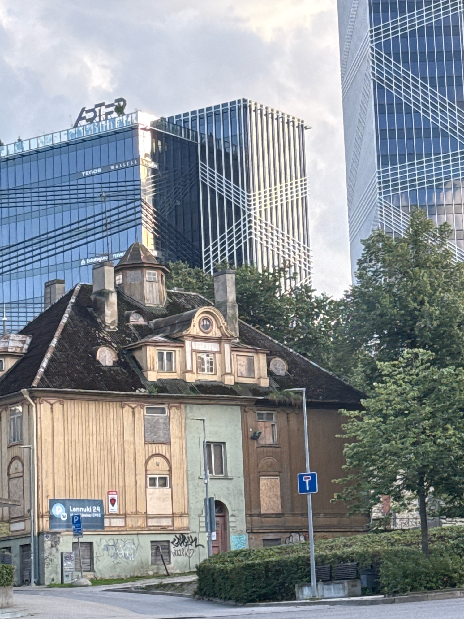 The image captures a street view in Tallinn, Estonia, showcasing a juxtaposition of old and new architecture. In the foreground, an aged, multi-story building with a dark, weathered roof and light-colored wooden siding stands prominently. This structure exhibits signs of disrepair, with boarded-up windows and peeling paint. Several architectural details, such as dormer windows and ornate trim, are visible on its facade. The building is adorned with graffiti on its lower level and a large blue and white sign that reads "Lennuki 24" with a parking symbol.
Behind the older building, two modern, imposing skyscrapers dominate the skyline. These glass-fronted structures feature vertical lines and reflective surfaces, indicating a contemporary urban setting. Lush green trees are interspersed between the buildings, softening the urban landscape. The sky is overcast, with hints of sunlight breaking through the clouds. A street sign with a blue background and a white symbol indicating a dead end or no through road is positioned near the foreground building. The overall scene suggests a location where historical architecture is being preserved or has been left to decay amidst rapid urban development.