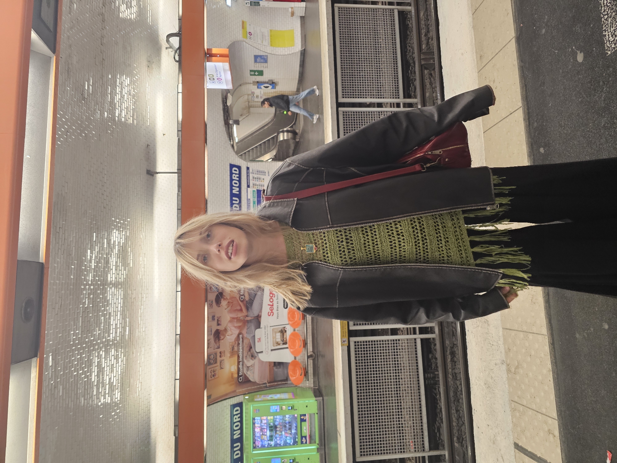 A young woman with long blonde hair stands on a subway platform, facing the camera with a neutral to slightly smiling expression. She is wearing a black leather-like jacket over a green crocheted top with distinctive fringe details, paired with dark pants. A red shoulder bag is visible on her left side.
The setting is a well-lit indoor transit station, identifiable as Gare du Nord in Paris due to prominent "DU NORD" signs on the walls. The station features light-colored tiled walls, orange architectural beams, and metal grates along the platform edge. In the background, another individual is seen ascending an escalator. The platform has various advertising posters, including one for "SeLoger" with the text "Q Studio indépendant" and "Vous êtes au bon". A green vending machine and another with orange dispensers are also visible on the platform. The overall lighting suggests it is daytime. The woman appears to be posing for a photograph while at the station.