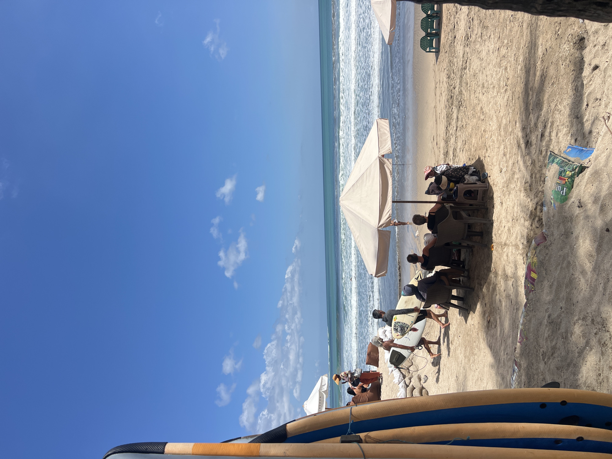 The image depicts a sunny day on Kuta Beach, Bali, Indonesia. In the foreground, several surfboards are leaning against each other, positioned on the sandy beach. Beyond the surfboards, a group of people are gathered under a large, light-colored umbrella, seated in chairs on the sand. Some are engaged in conversation, while others appear relaxed. Two individuals are walking towards the ocean, each carrying a surfboard. The ocean stretches out to the horizon, with gentle waves breaking on the shore. The sky is a vibrant blue, dotted with a few scattered white clouds. Further down the beach, other umbrellas and beach chairs are visible, suggesting a lively beach atmosphere. The scene captures a typical day at a popular Indonesian surf spot, with a blend of beachgoers and surfers enjoying the coastal environment.