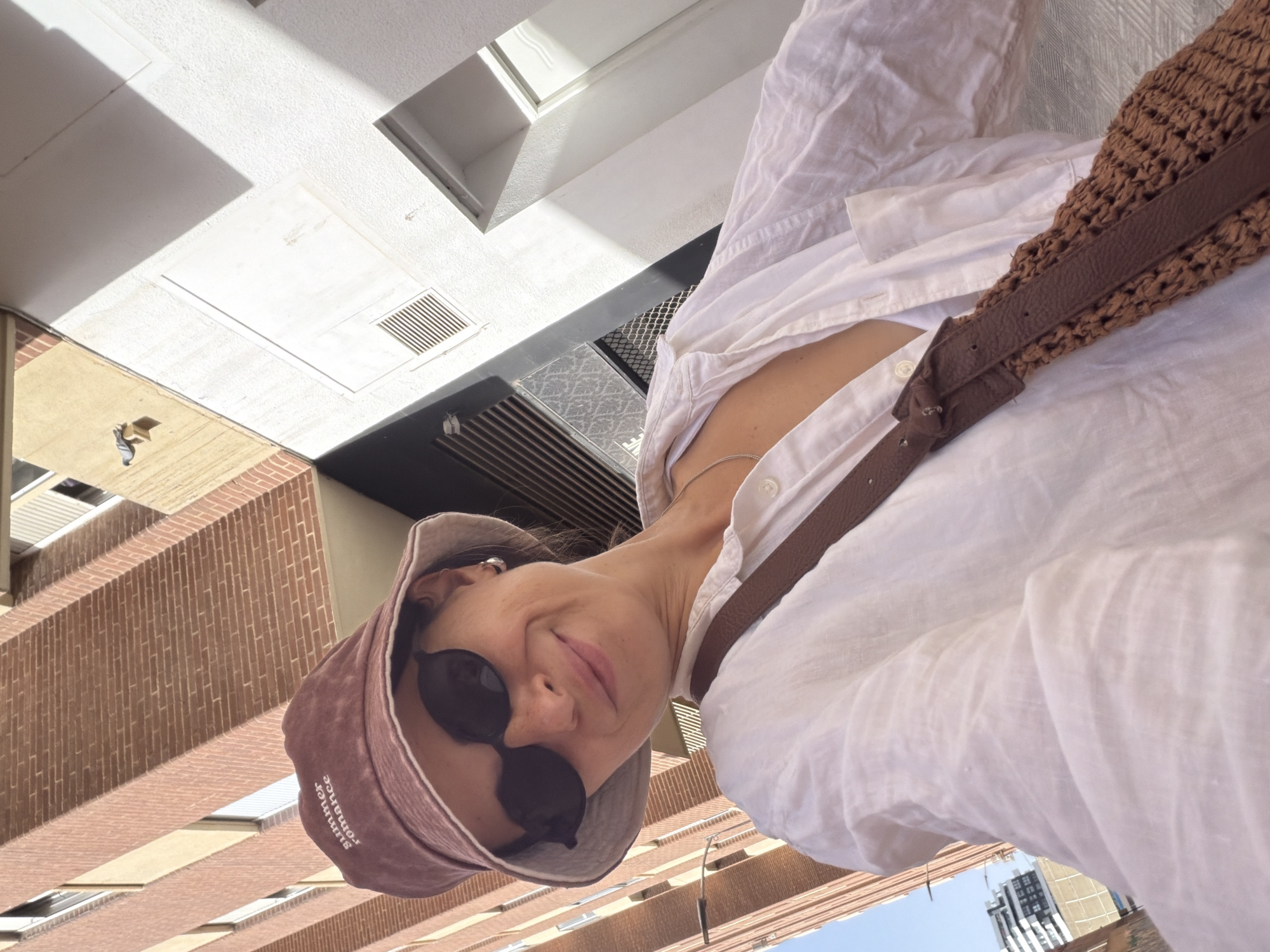 A woman takes a selfie in an urban environment. She is wearing a bucket hat that reads "summer romance", sunglasses, and a white button-down shirt. A brown strap from a bag is visible across her chest. The background consists of a brick building and a white wall with shadows cast by sunlight.