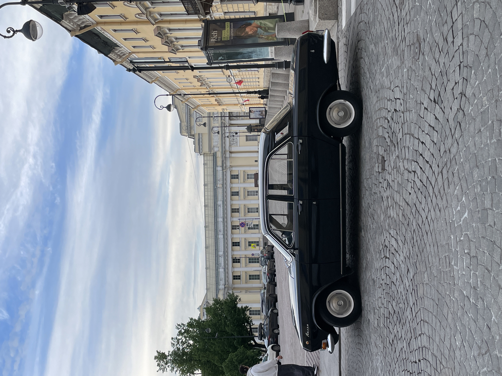 A classic black car, identified as a GAZ-21 Volga by the Cyrillic text "Волга" on its side, is parked on a grey cobblestone street in Saint Petersburg, Russia. The car, with its chrome details, is positioned on the right side of the frame, facing away from the viewer.
The street is paved with intricately arranged cobblestones. On the left, a row of stately, pale yellow buildings with classical architectural features like columns, cornices, and decorative windows stretches into the distance, indicative of Saint Petersburg's historic urban landscape.
The sky above is bright blue with wispy white clouds, suggesting clear daytime weather. Several ornate black street lamps are visible, complementing the historic feel of the street. Green trees add natural elements to the scene.
Further down the street, a line of more contemporary cars is parked. On the sidewalk near the vintage car's rear, a person in a white hoodie and dark pants walks, looking downwards.
An advertising billboard is visible on the left side of the street, displaying the English word "Rich" and some Cyrillic text below it, which appears to read "ОДЕЖДА КУЛЬТУРНАЯ БЕЗ ЦЕРЕМОНИЙ" ("Cultural Clothes Without Ceremonies" or "Informal Attire").
The scene captures a peaceful daytime moment in a historic city, featuring a blend of vintage and modern elements.