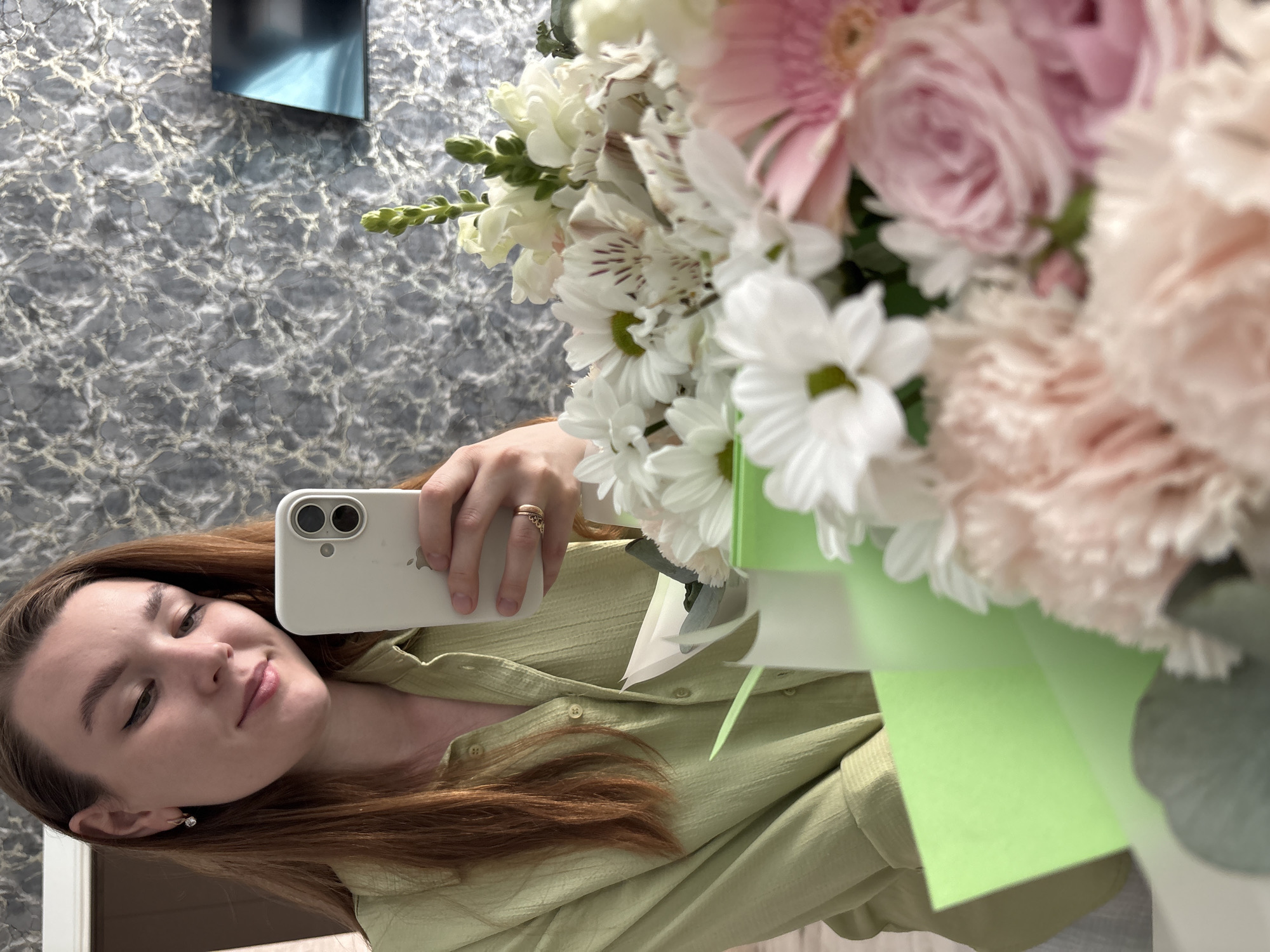 A woman is taking a selfie with a bouquet of flowers. She is holding a white smartphone. The flowers are a mix of pink, white, and green. The background is a grey marbled wall with a dark rectangle hanging on it. The city is Izhevsk, Russia.